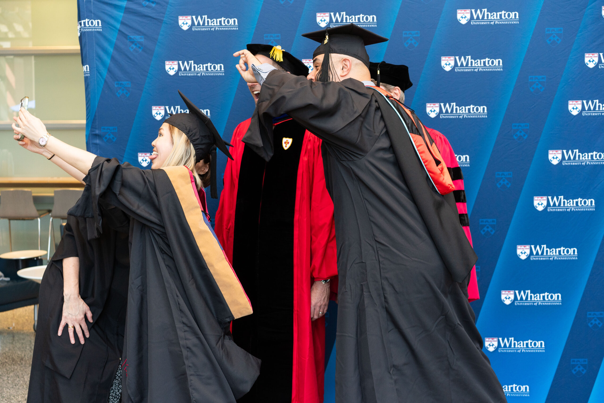 Graduates wearing caps and gowns take a selfie at a commencement ceremony in front of a Wharton School, University of Pennsylvania backdrop.