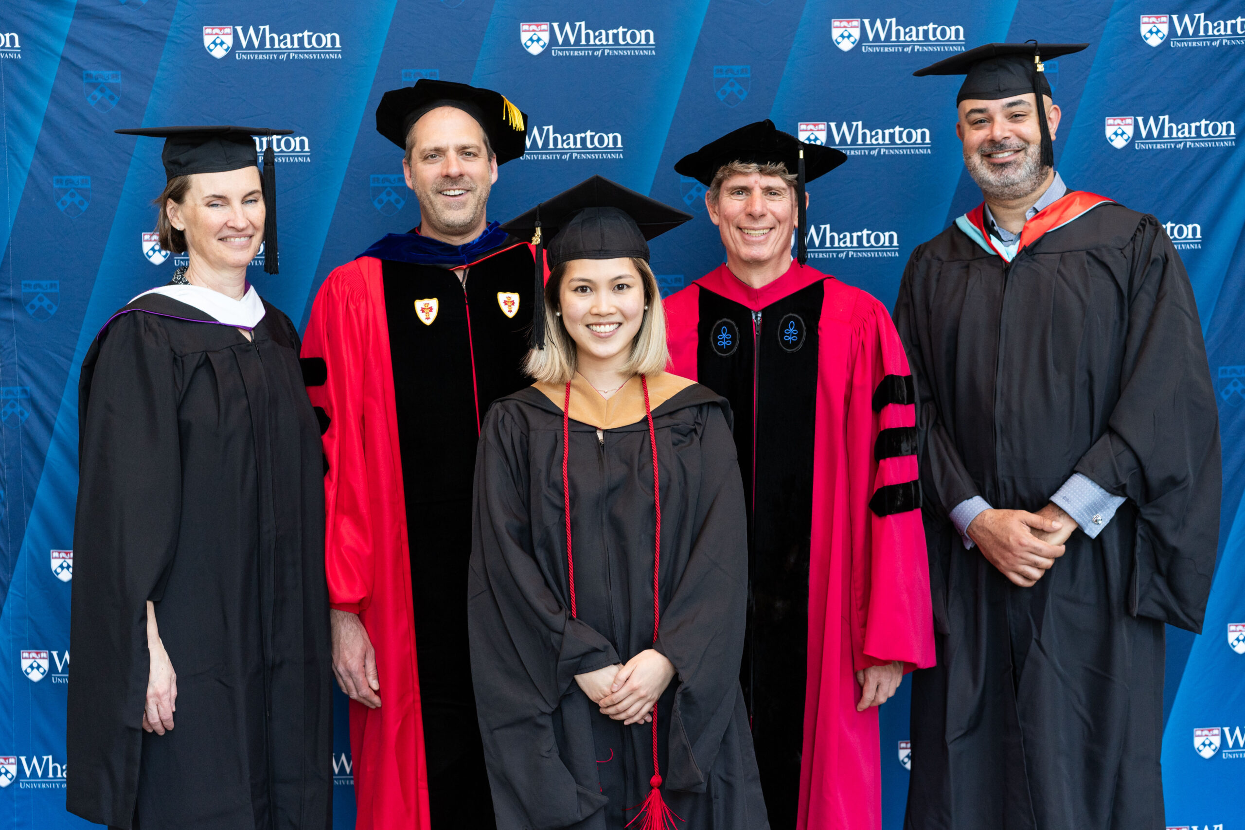 A small group of people in academic regalia, possibly at a graduation ceremony, standing in front of a backdrop with "Wharton University of Pennsylvania" logos.