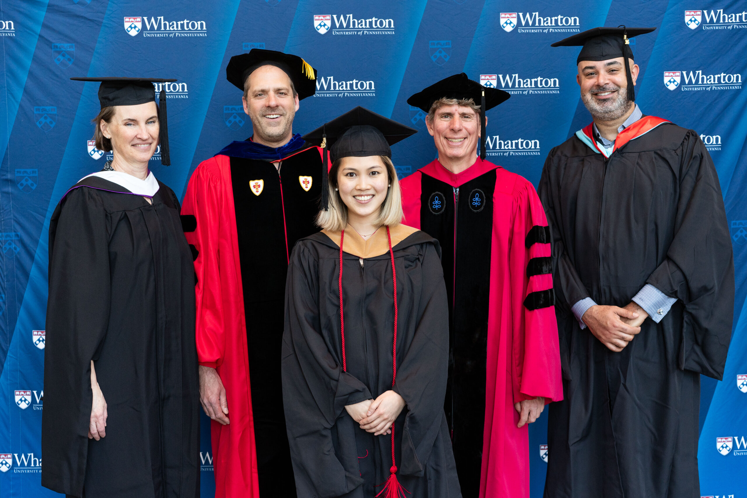 A small group of people wearing academic regalia, including graduation gowns and caps, standing in front of a Wharton School of the University of Pennsylvania backdrop.