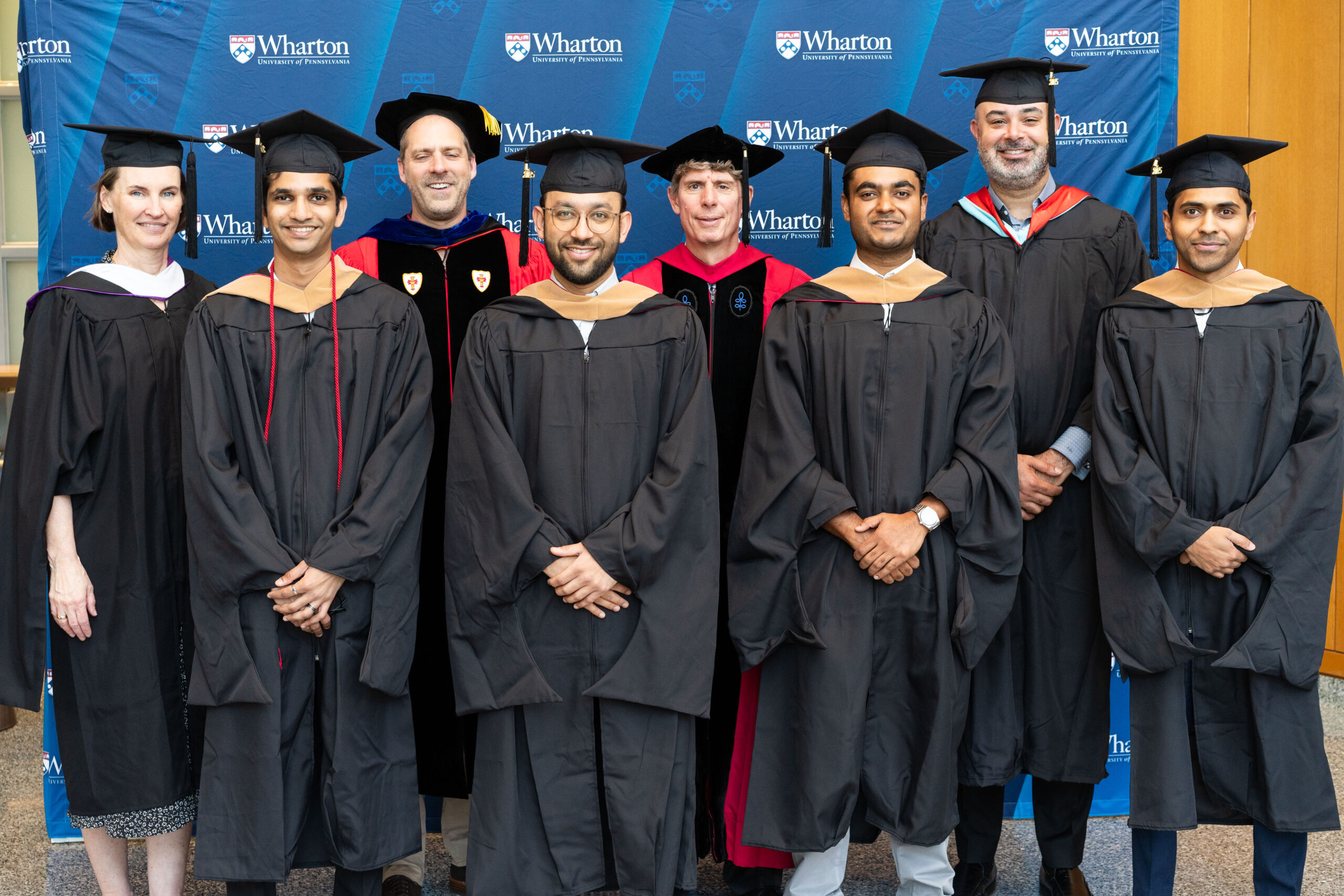 A group of people in graduation attire, including caps and gowns, posing in front of a Wharton School background, possibly representing a graduation ceremony.