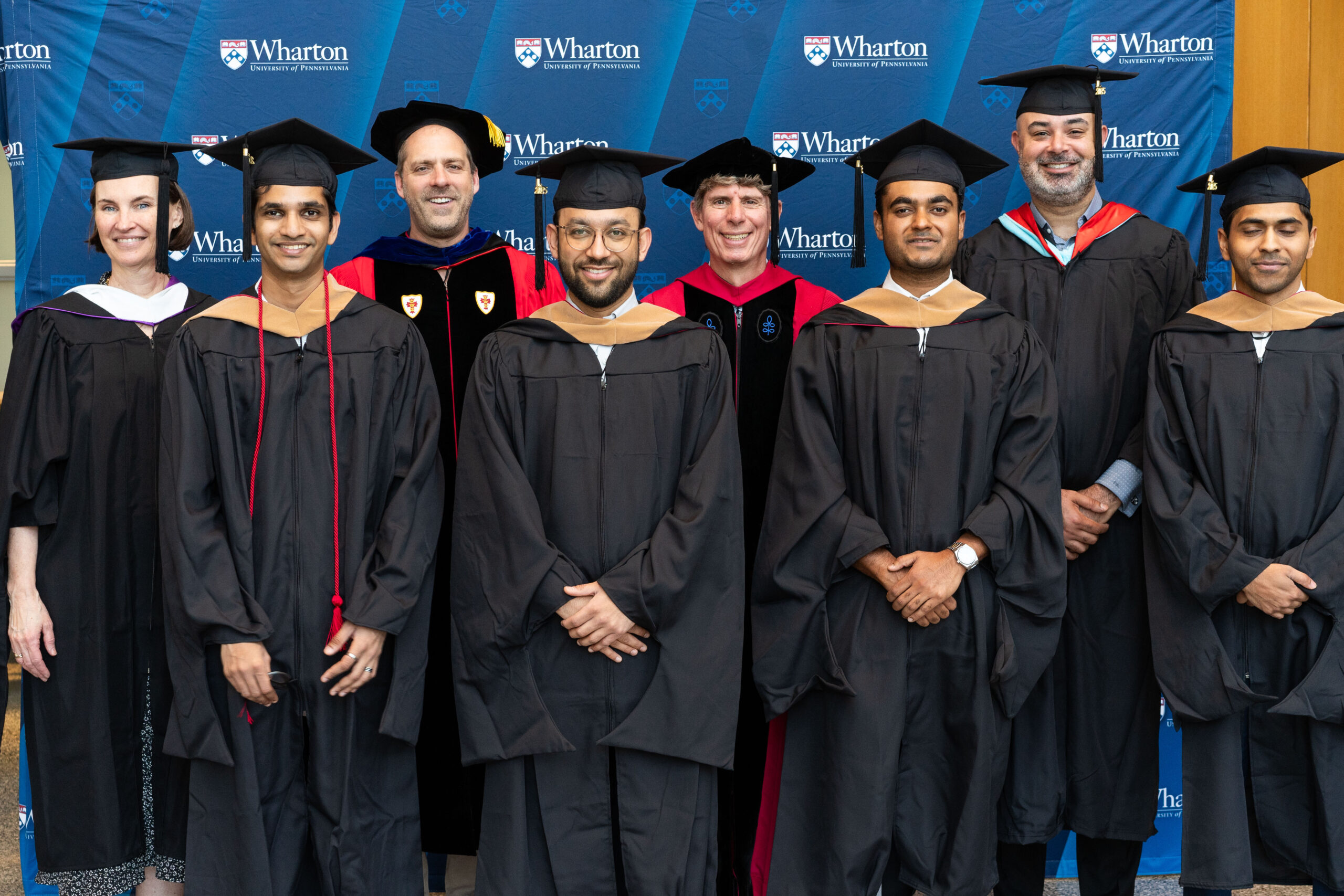 A group of graduates in caps and gowns stand with faculty members in academic regalia, in front of a backdrop displaying the Wharton School logo.