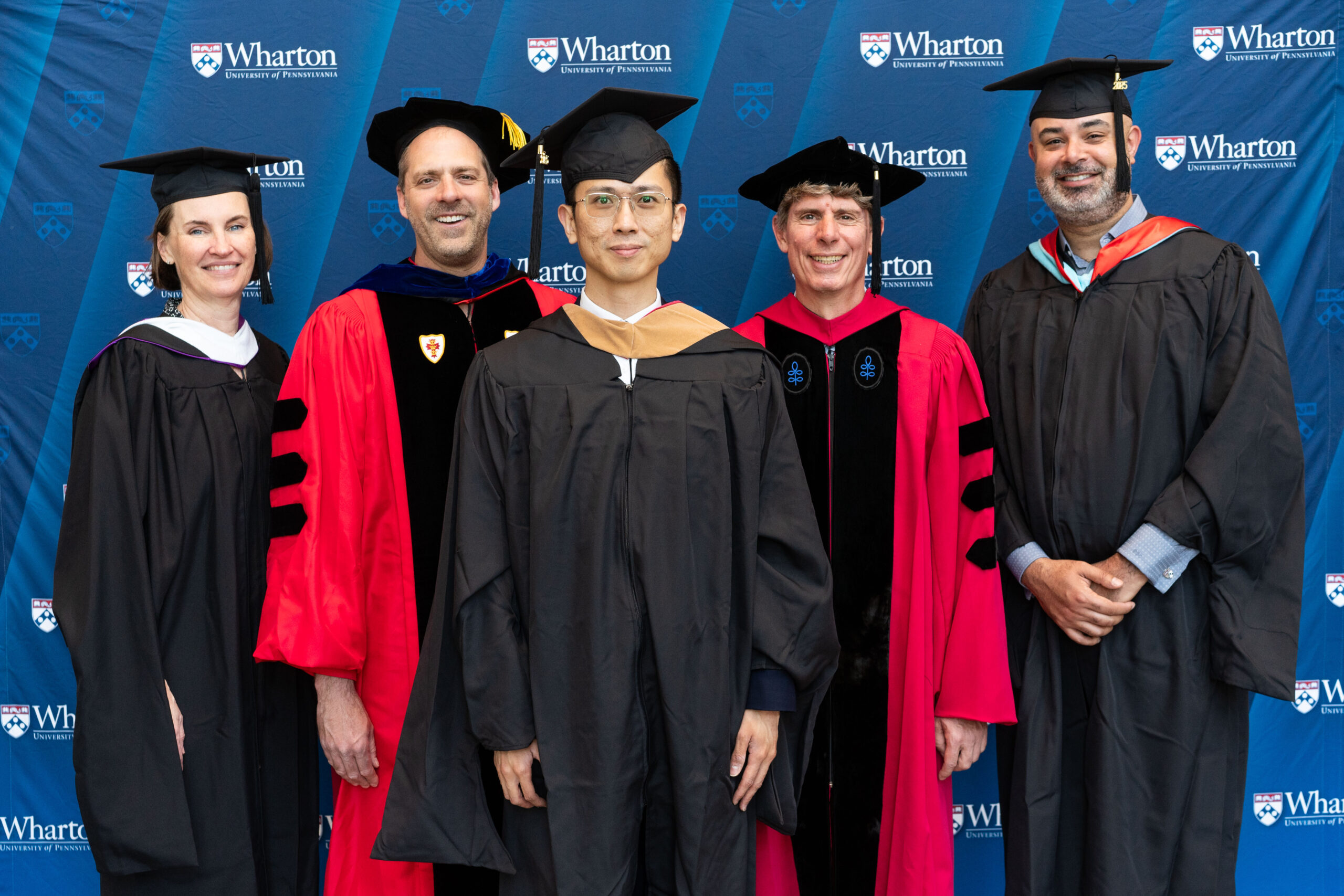 A group of people in graduation attire are standing in front of a Wharton School backdrop. They are wearing caps and gowns, indicating a graduation or academic event.