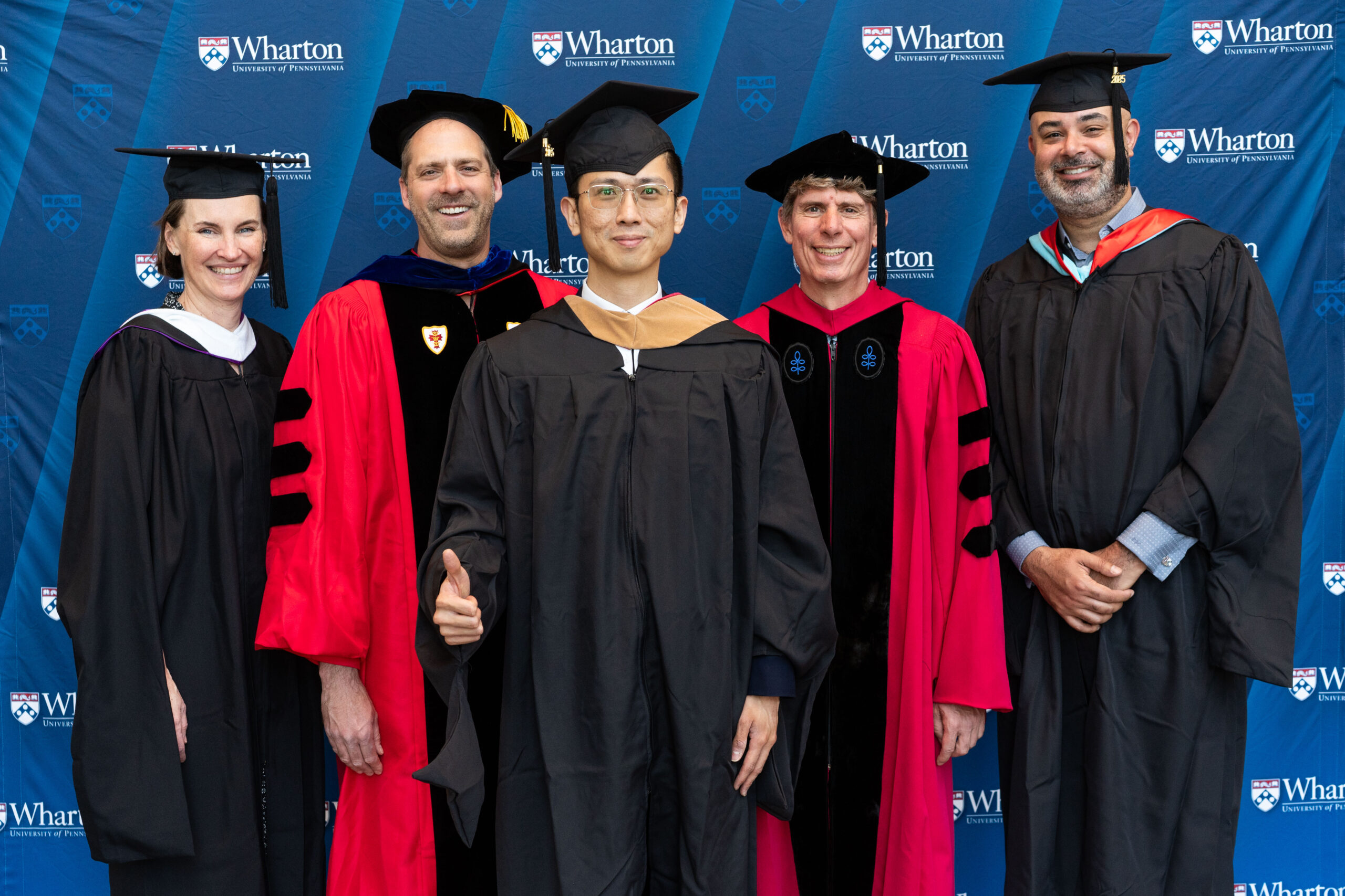 A group of people in academic regalia, possibly graduates and faculty, standing in front of a Wharton School of the University of Pennsylvania backdrop. One person is giving a thumbs-up.
