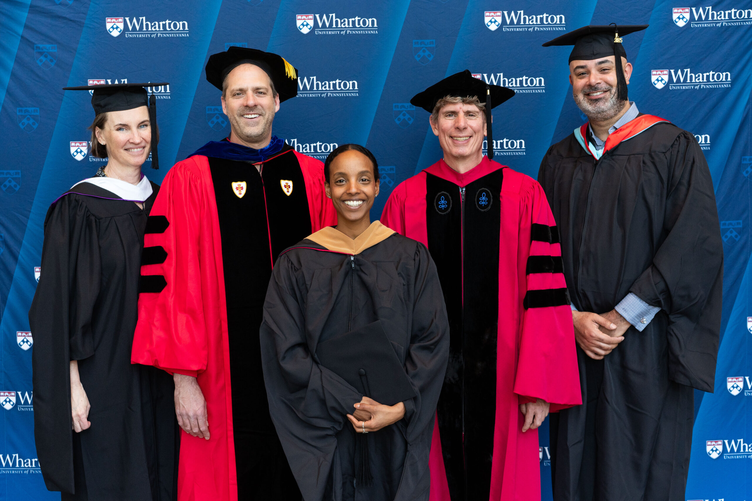 A small group of people in academic gowns and caps, posing in front of a backdrop with the Wharton School logo. The group appears to be at a graduation or academic event.