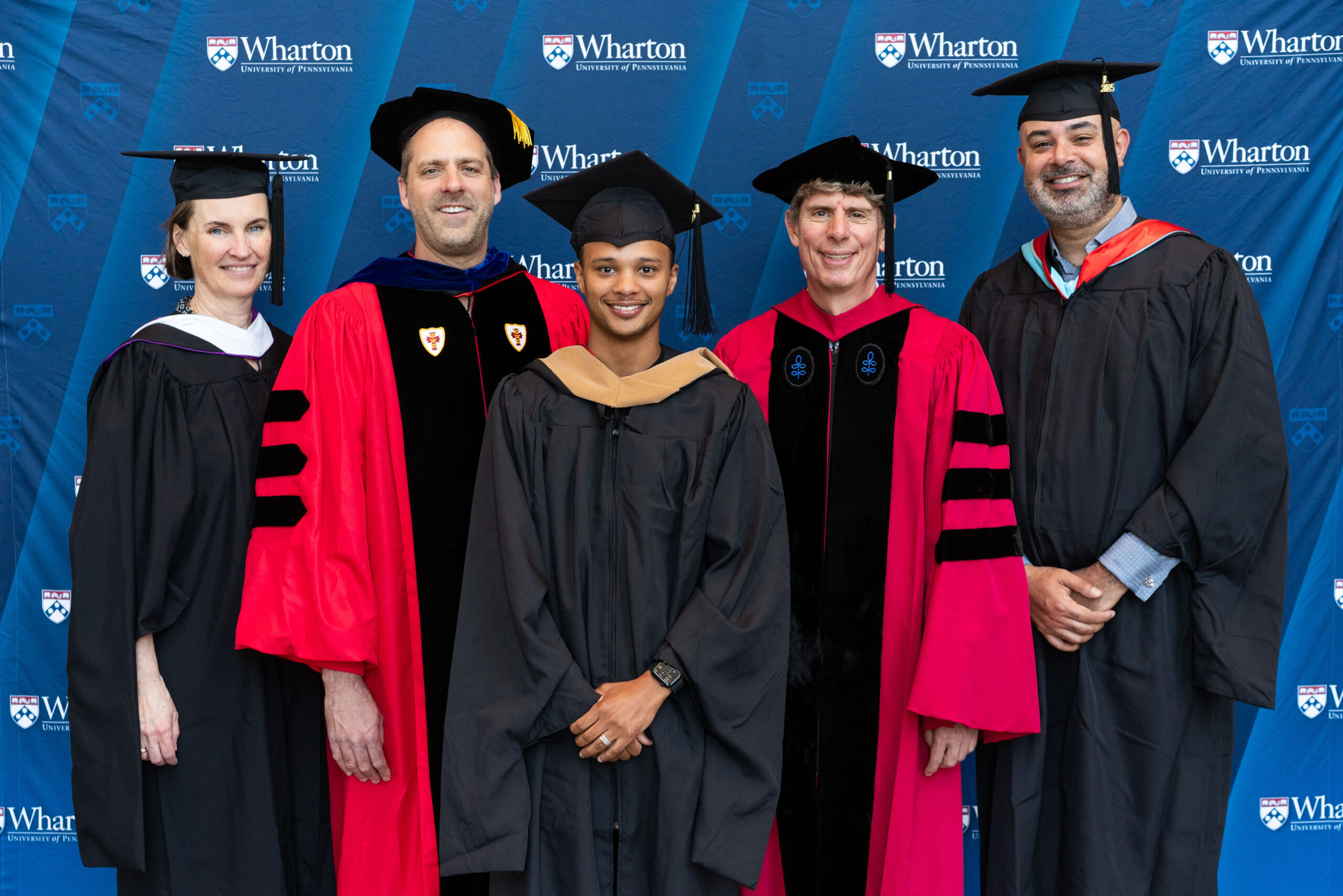A small group of people in graduation attire, including robes and caps, posing in front of a backdrop with the Wharton School logo.