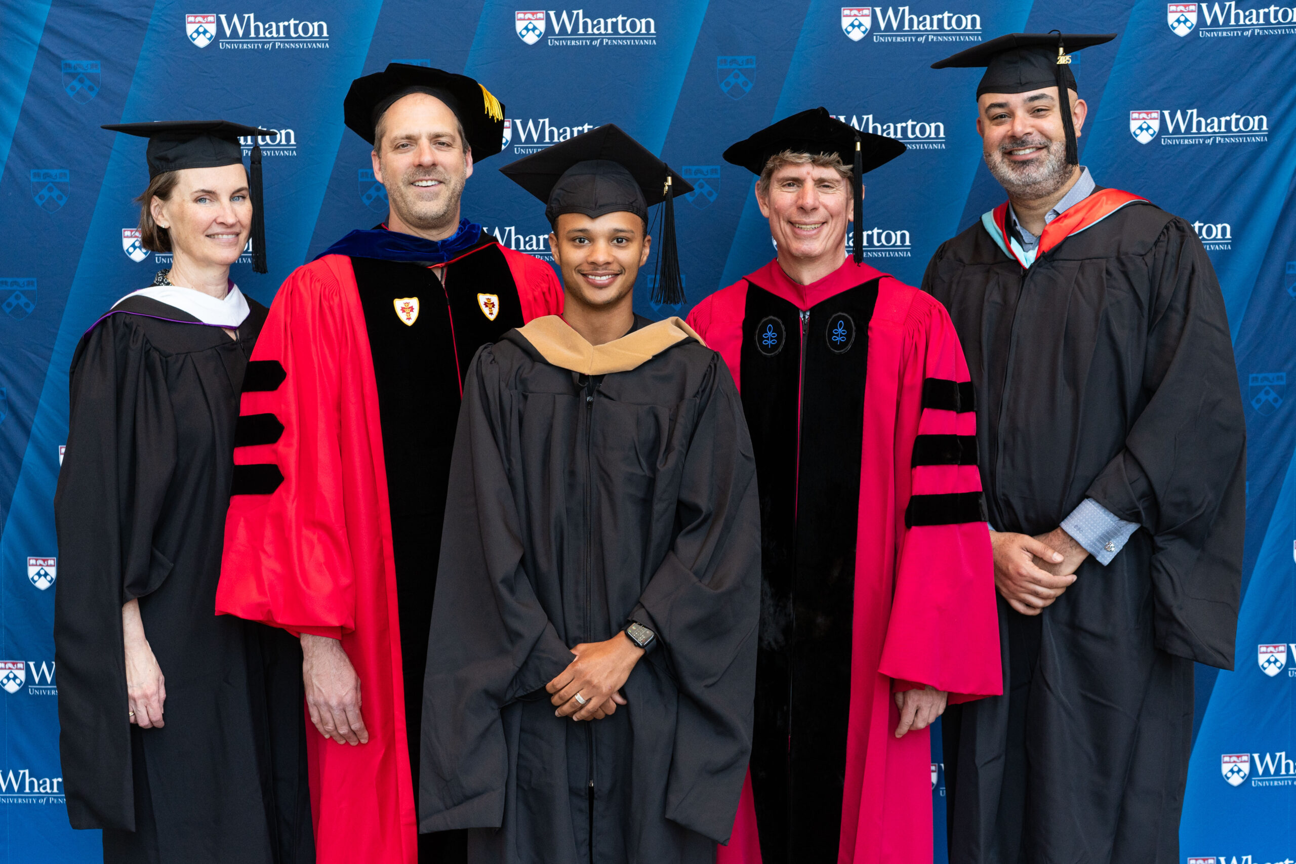 A group of individuals in academic regalia posing for a graduation photo against a Wharton School of the University of Pennsylvania backdrop.