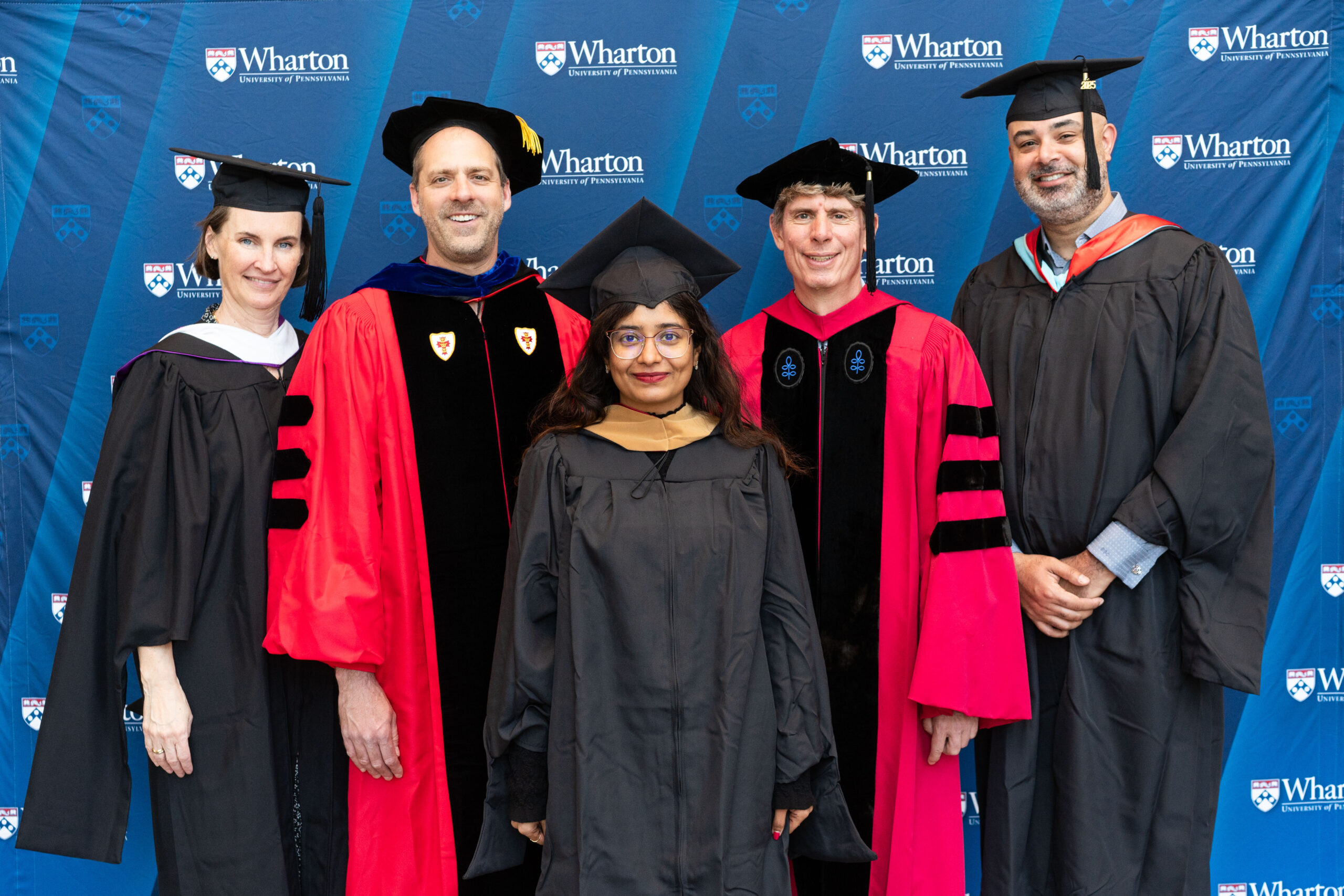A small group of people dressed in academic regalia, posing in front of a Wharton School backdrop. This represents a graduation or academic ceremony.