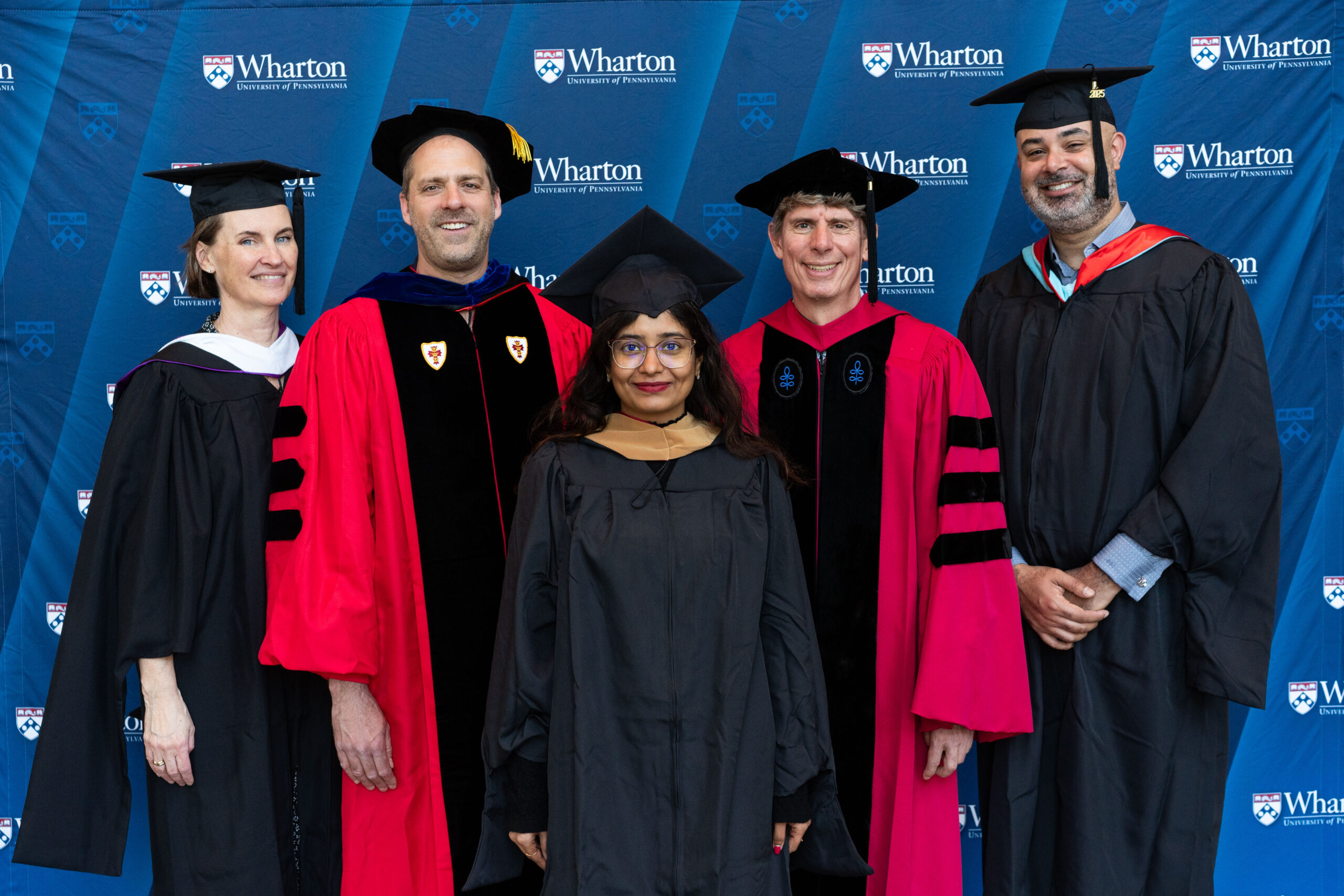 A group of people in academic regalia stand against a backdrop featuring the University of Pennsylvania Wharton School logo, suggesting a graduation or academic event.