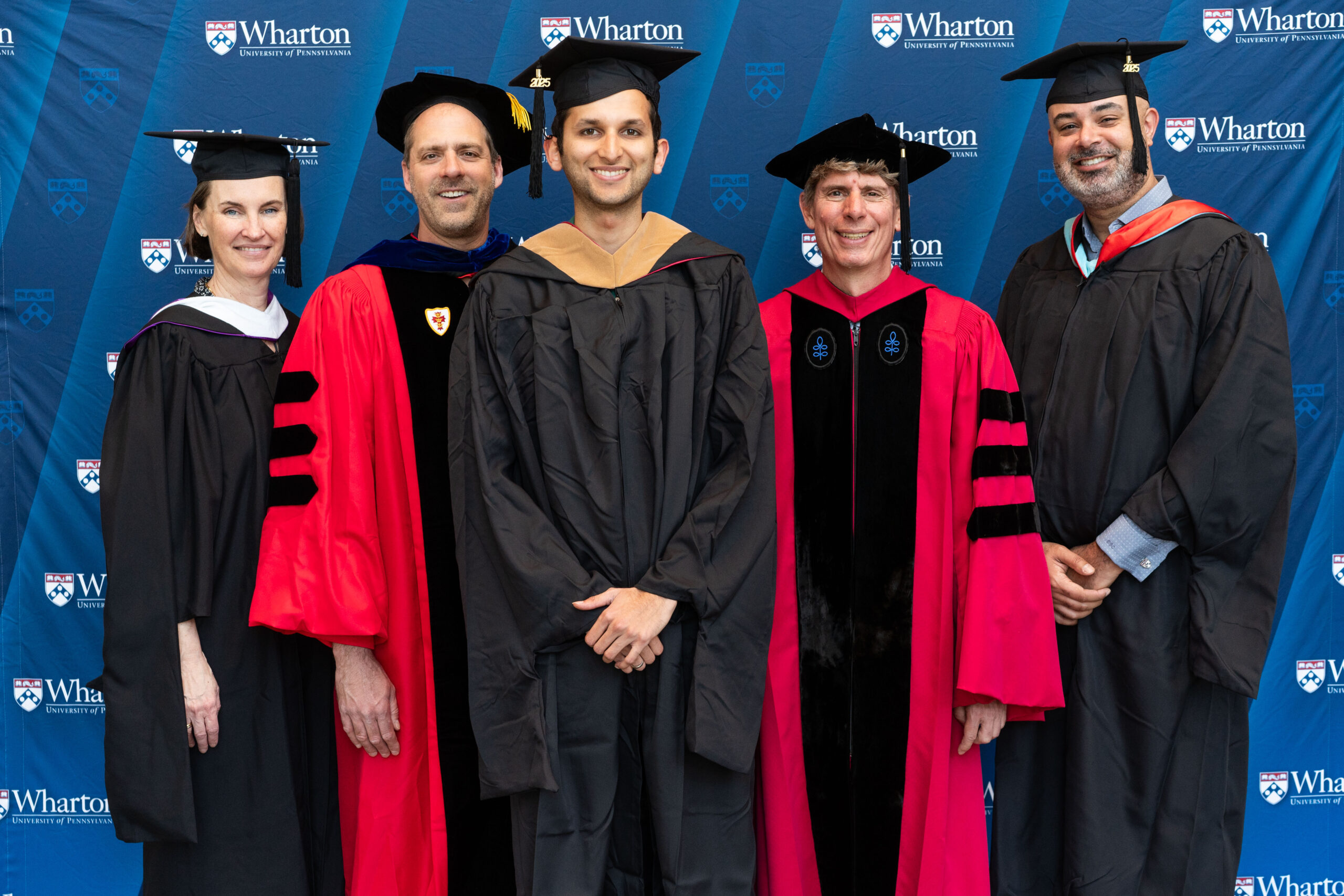 A group of people in academic regalia posing in front of a Wharton School backdrop, likely celebrating a graduation or academic event.