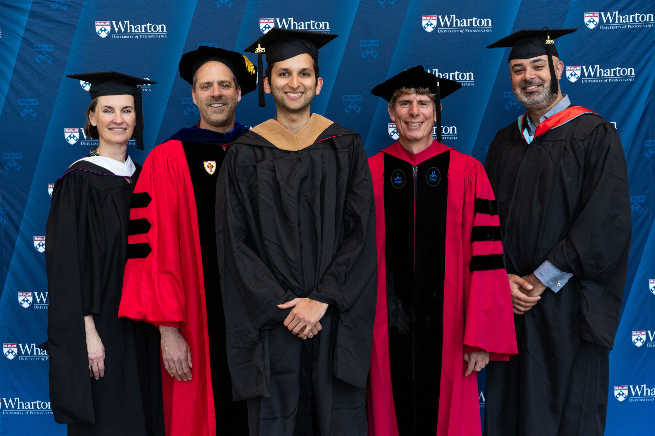 A group of people in academic gowns and caps stands in front of a Wharton School backdrop, suggesting a graduation or commencement event.