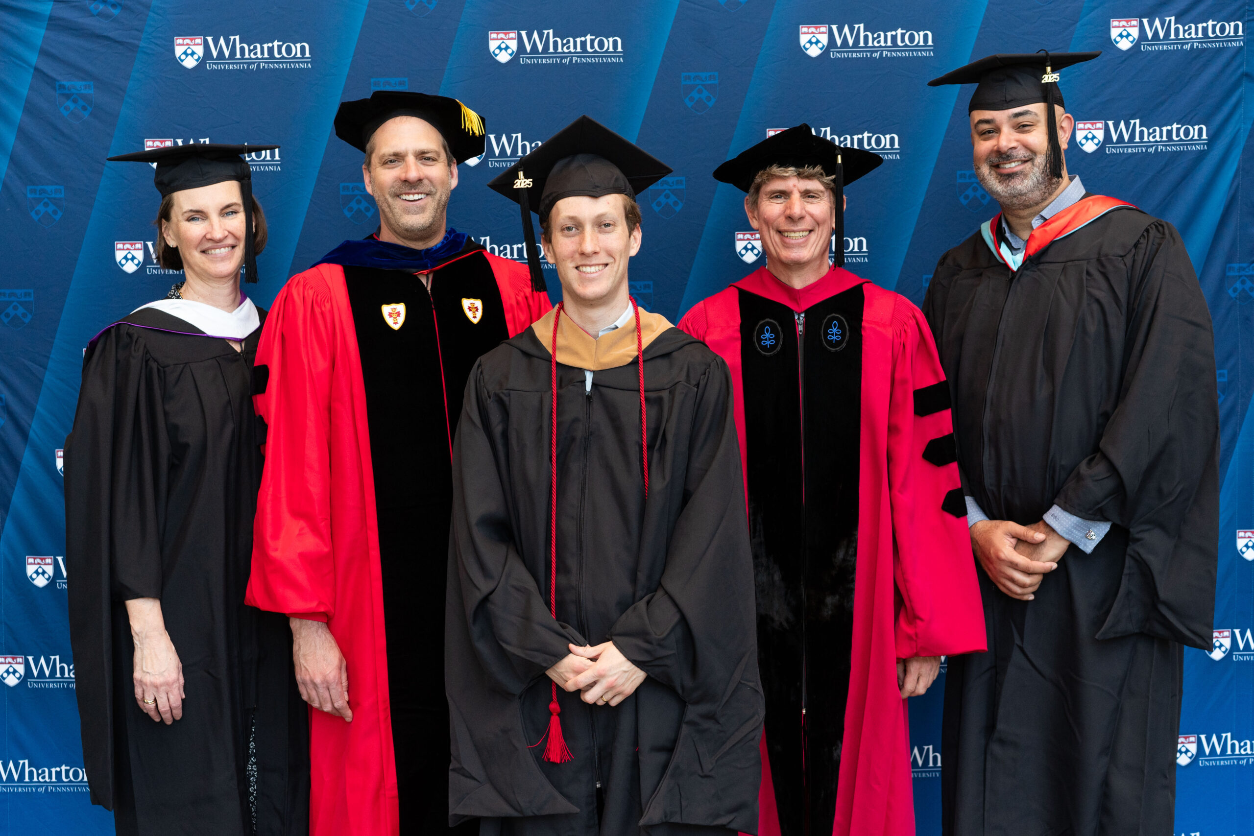 A group of people in academic regalia, including caps and gowns, standing in front of a backdrop with the Wharton School logo, suggesting a graduation or academic event.