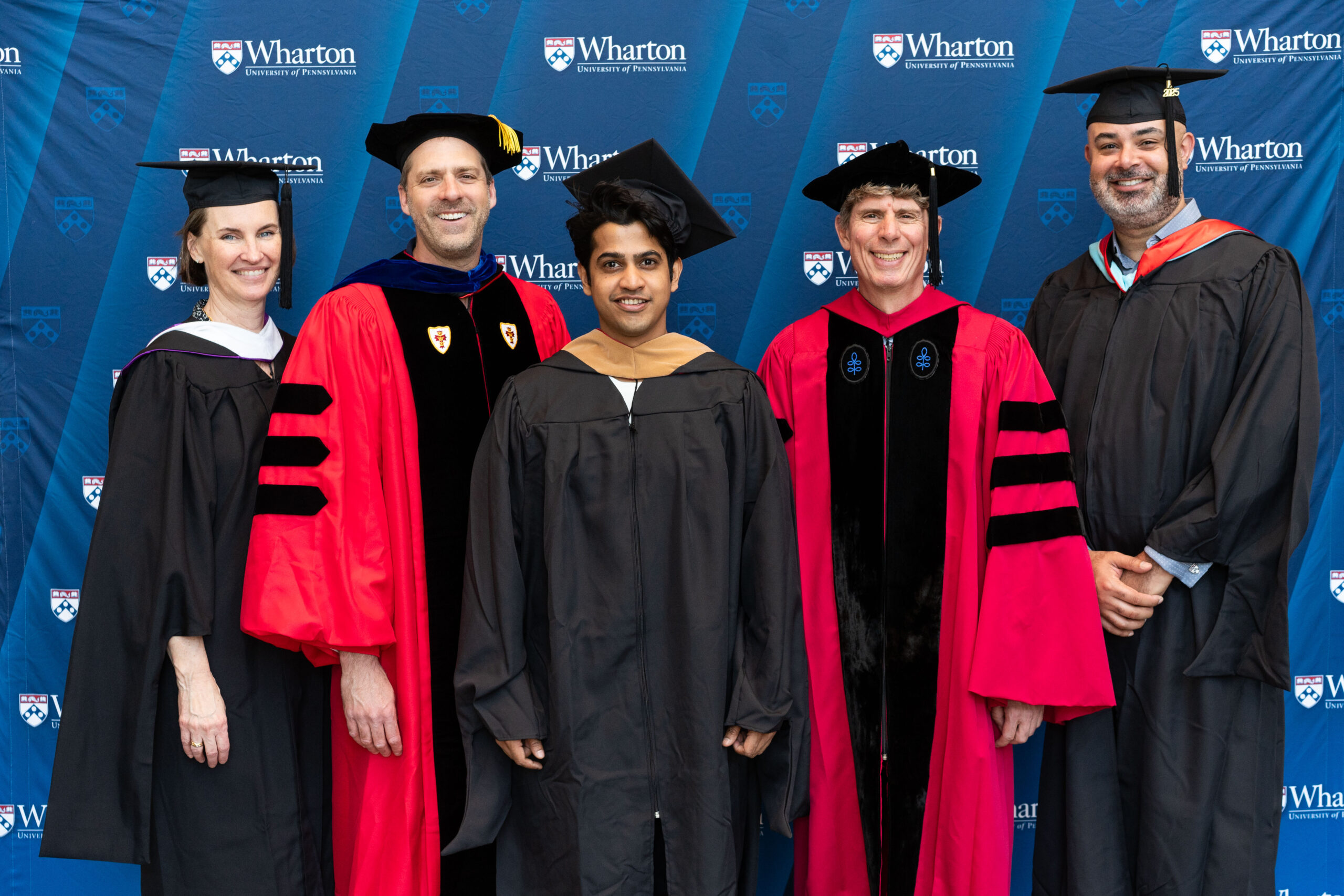 A group of people in academic regalia posing in front of a Wharton School backdrop, suggesting a graduation or academic event.