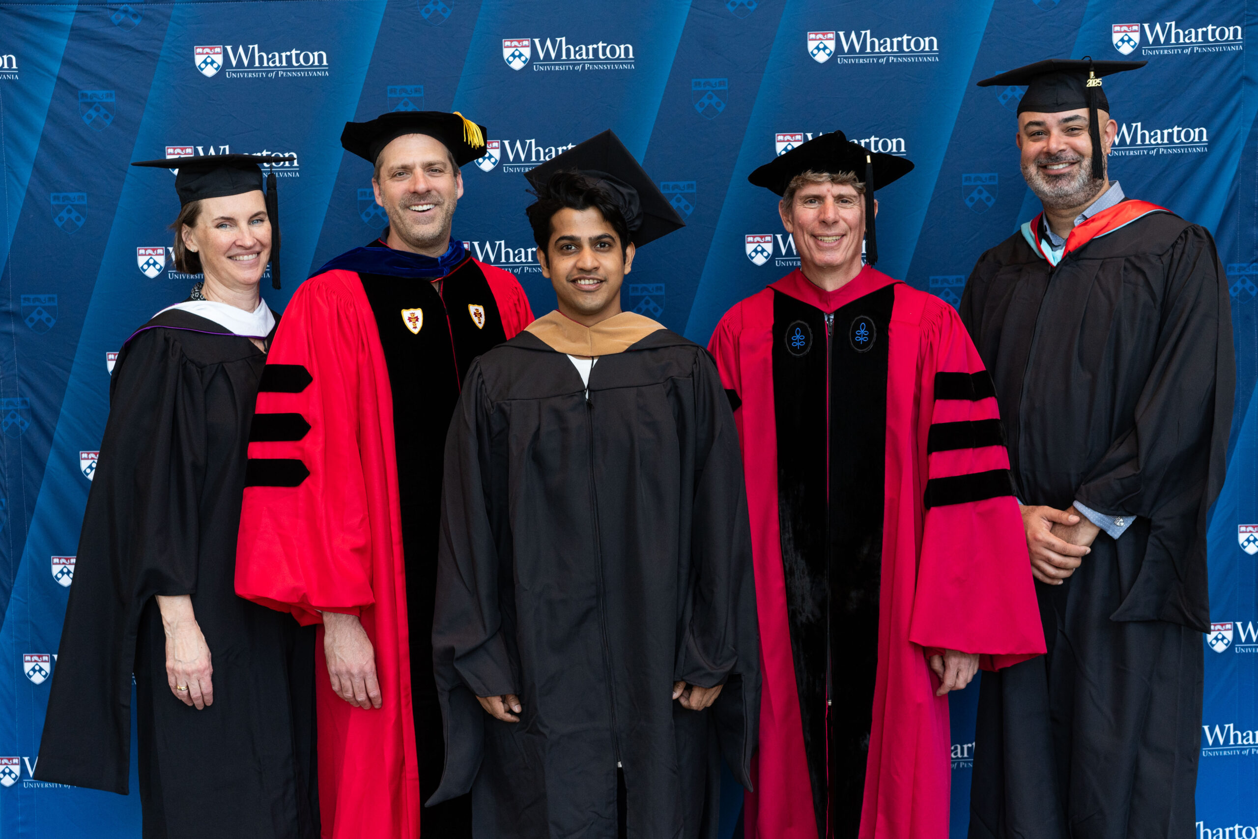 A group of people wearing academic regalia is posing in front of a Wharton School backdrop. Some are in graduation gowns, while others are in faculty robes with hoods.