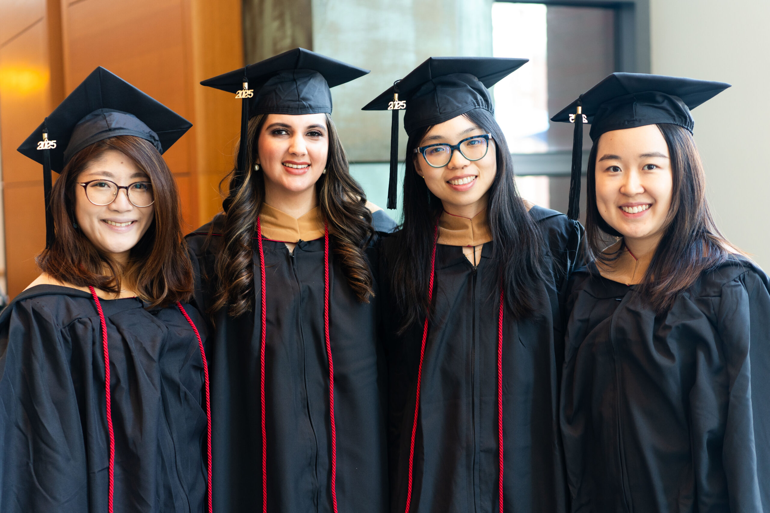 A small group of graduates in black caps and gowns, smiling and posing indoors. They wear tassels and red cords signifying academic achievements.