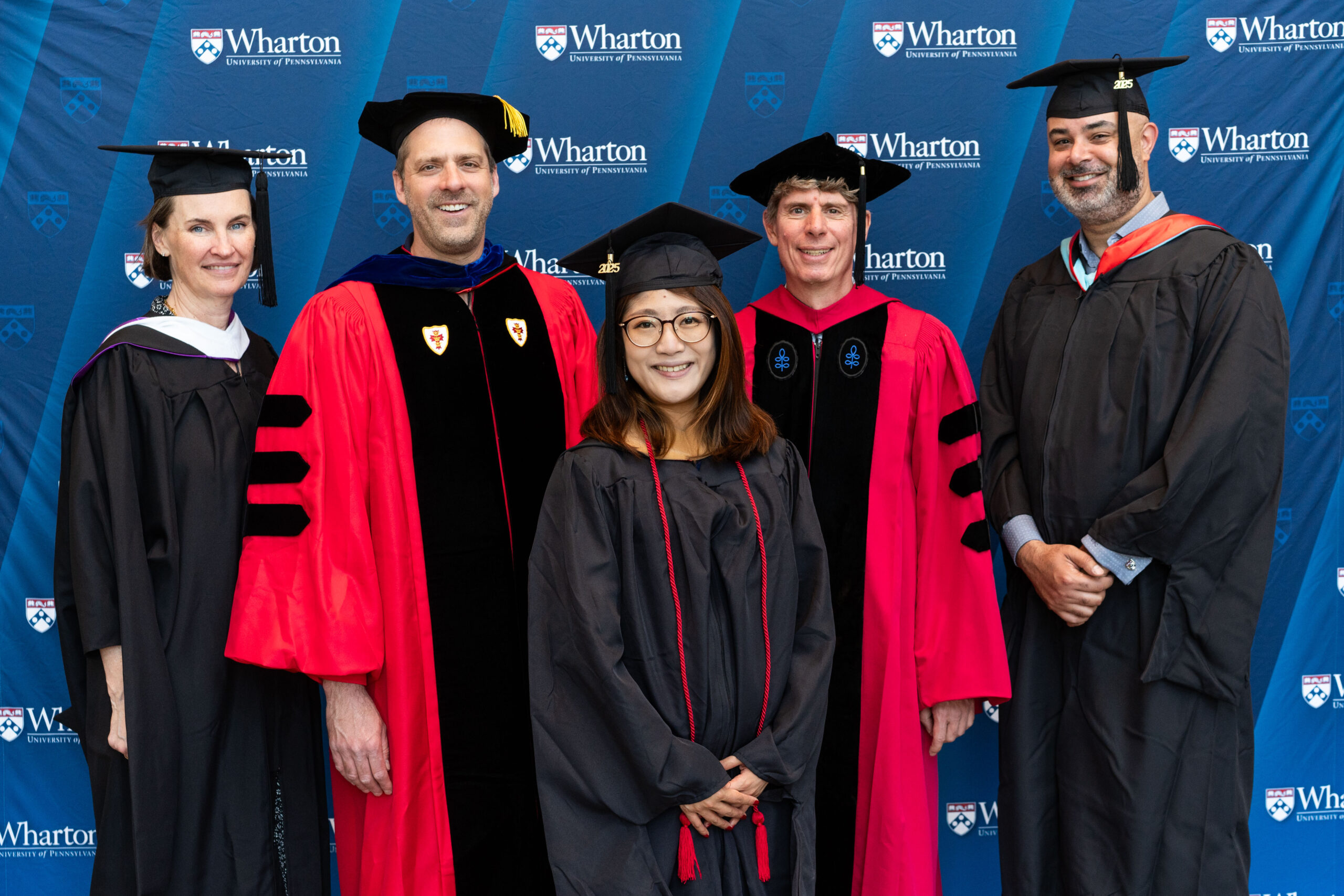 A group of people in academic regalia smiling in front of a Wharton School backdrop. The image likely represents a graduation or academic event.