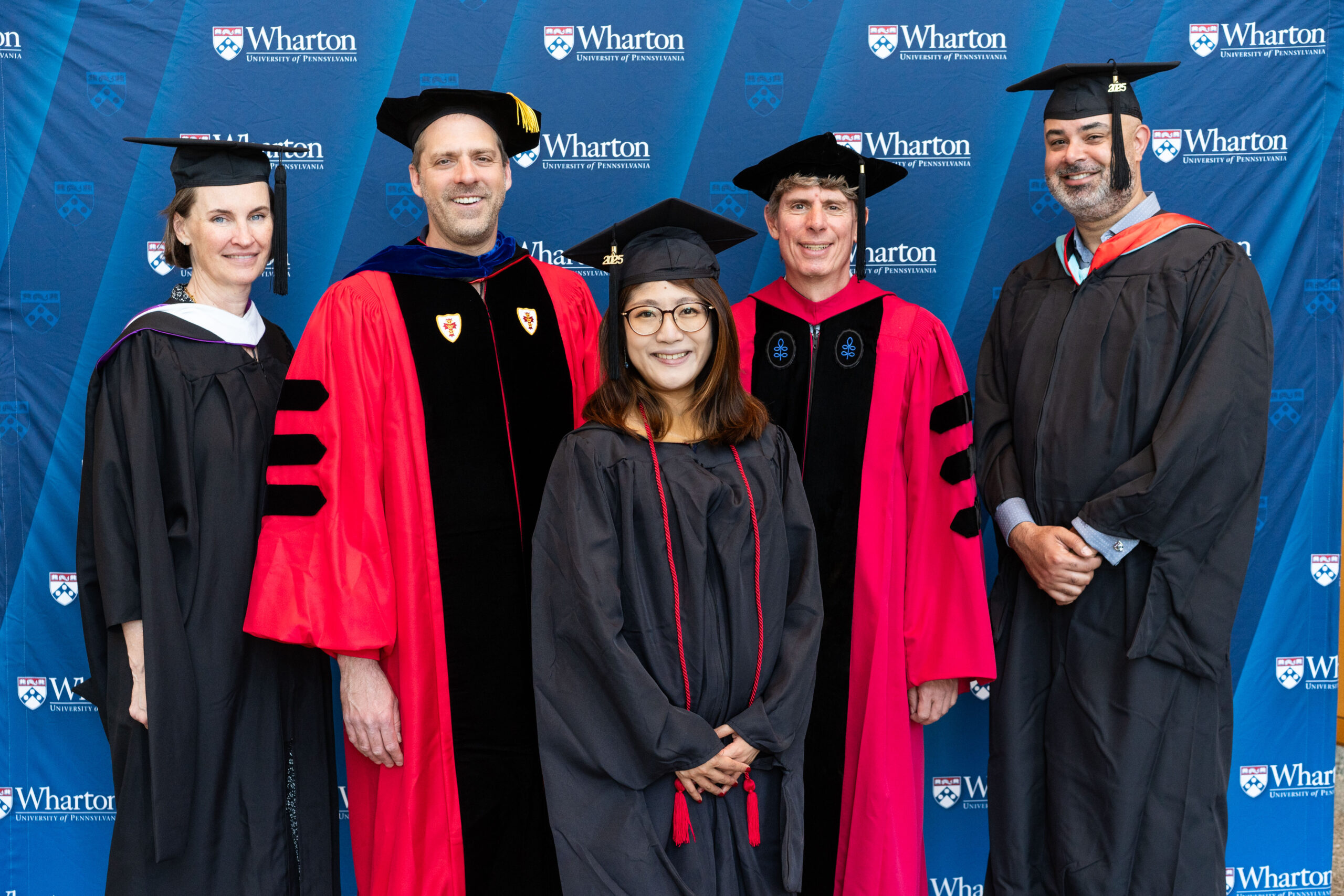 A small group in academic regalia, likely at a graduation event, with Wharton School branding in the background. They appear to be celebrating an academic achievement.