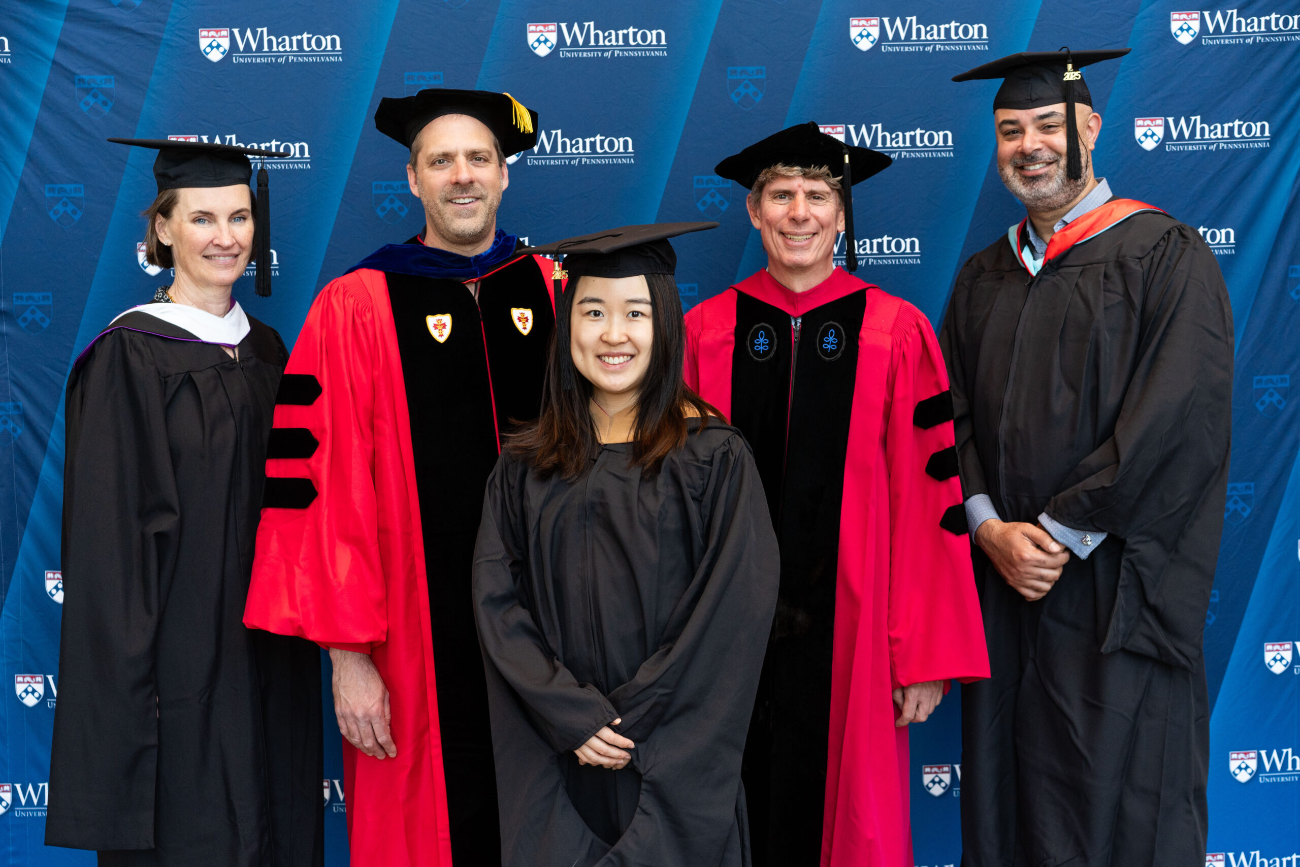 A group of people in academic regalia stand in front of a blue Wharton School backdrop, suggesting a graduation or academic event.