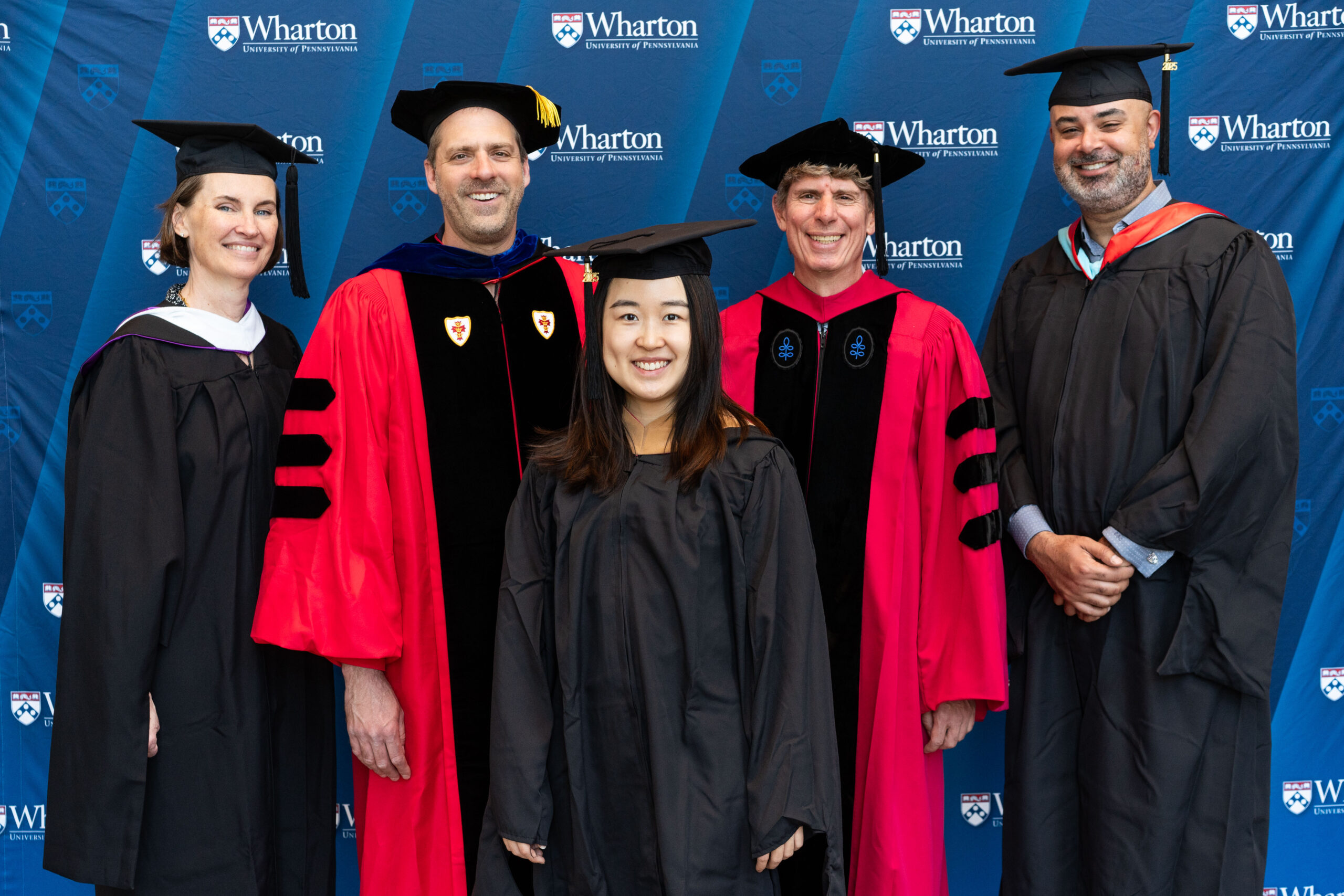 A group of people dressed in academic regalia, including caps and gowns, standing in front of a Wharton School of the University of Pennsylvania backdrop.