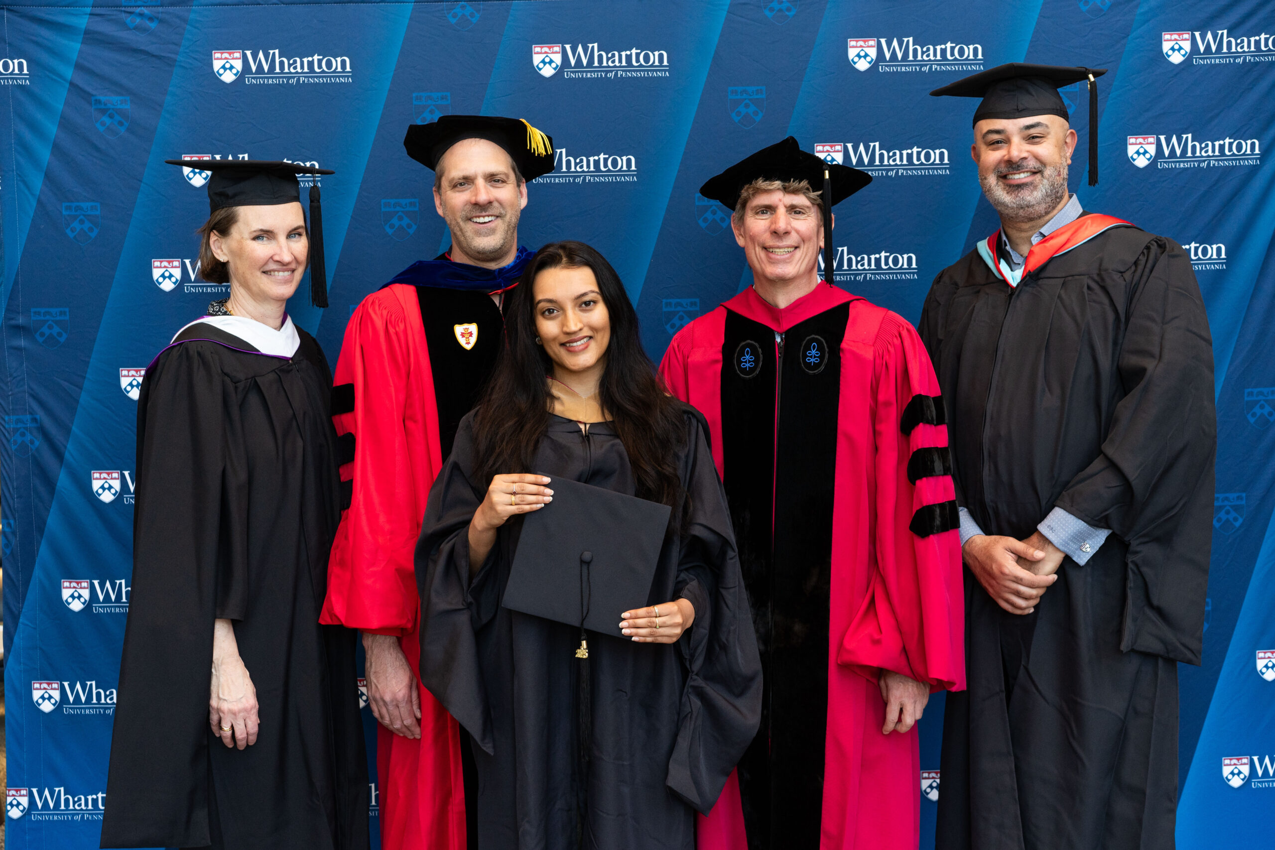 A group of individuals in academic regalia, including caps and gowns, standing in front of a Wharton School of the University of Pennsylvania backdrop. The group includes graduates and possibly faculty members.