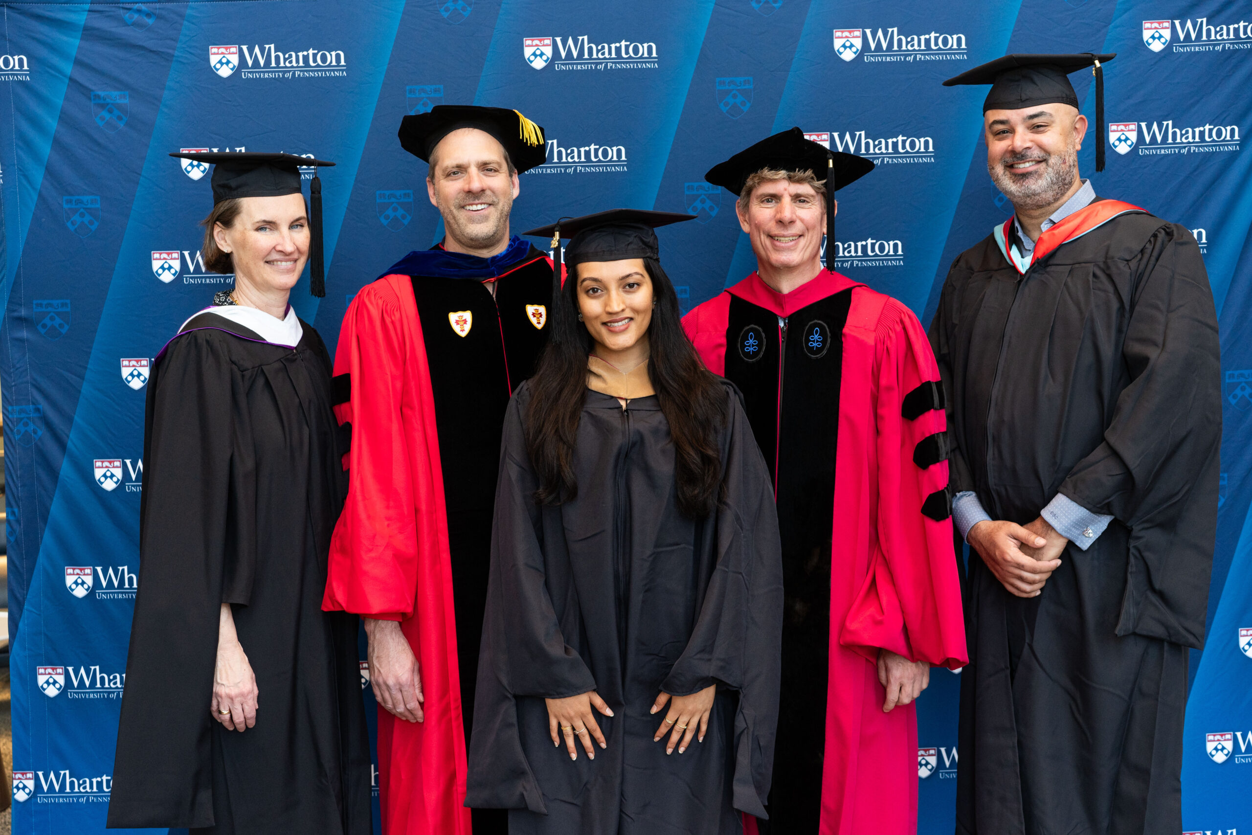 A group of people in graduation attire pose in front of a backdrop with the Wharton School logo. Some are wearing red academic robes and hats, while others are in black graduation gowns and caps.