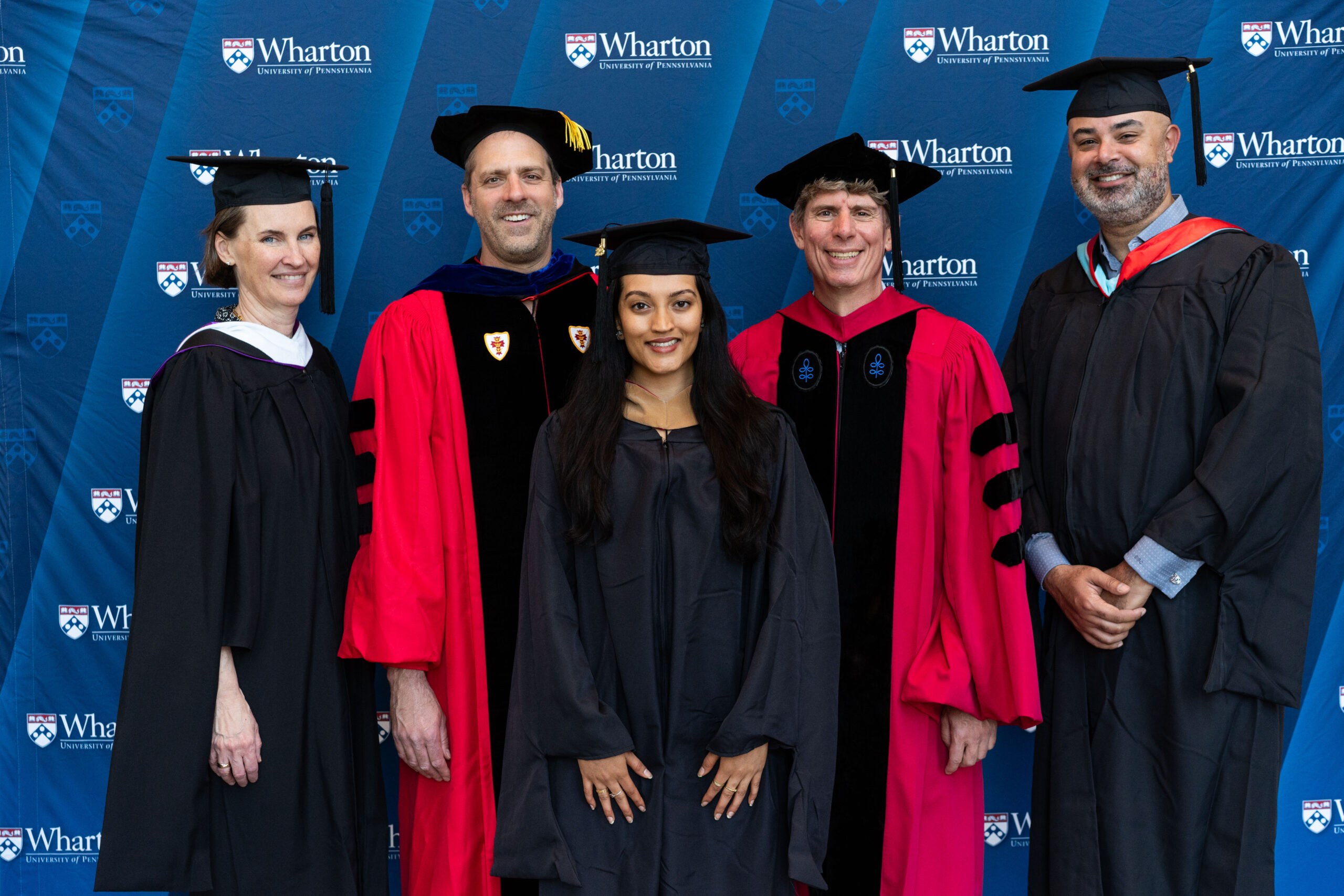 A small group in academic regalia, including gowns and caps, posing in front of a Wharton School backdrop, likely during a graduation event.