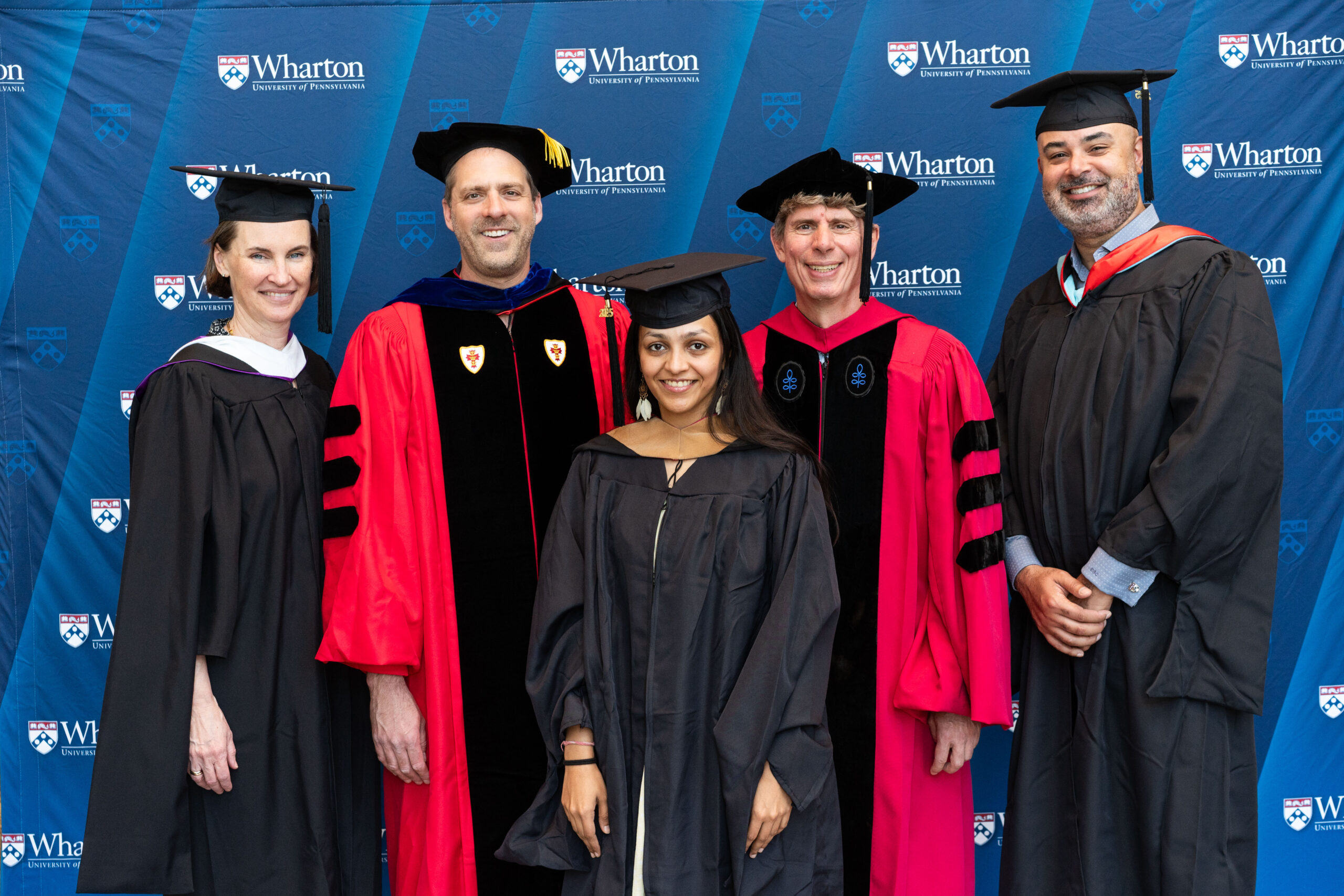 A group of individuals in academic regalia, including a graduate and faculty members, stand in front of a backdrop featuring the Wharton School of the University of Pennsylvania logo.