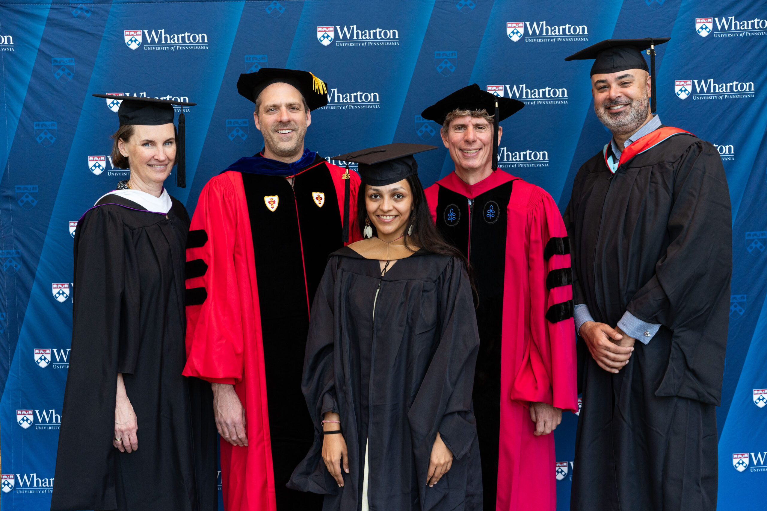 A group of people in academic regalia posing for a graduation photo in front of a backdrop with "Wharton University of Pennsylvania" logos.