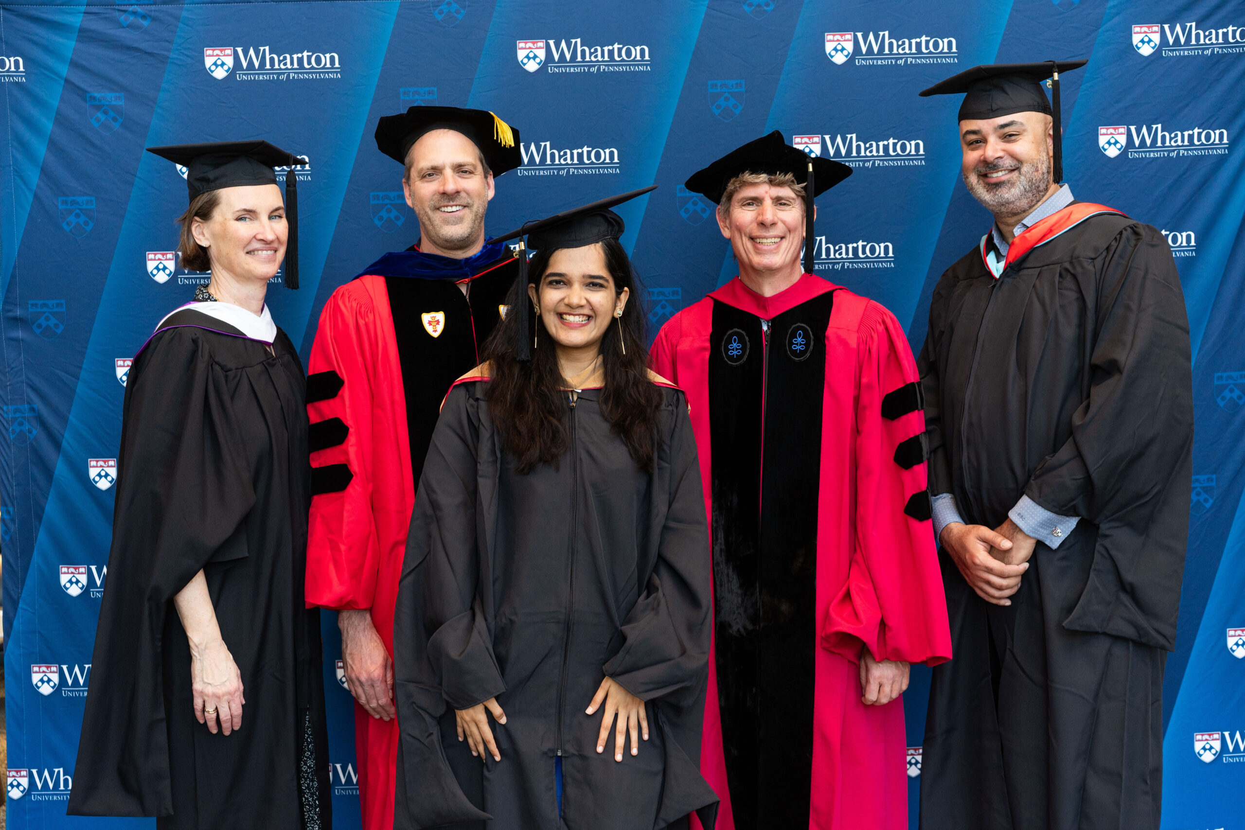 A group of people dressed in academic regalia, posing in front of a backdrop with the Wharton School of the University of Pennsylvania logo, signifying a graduation or academic event.