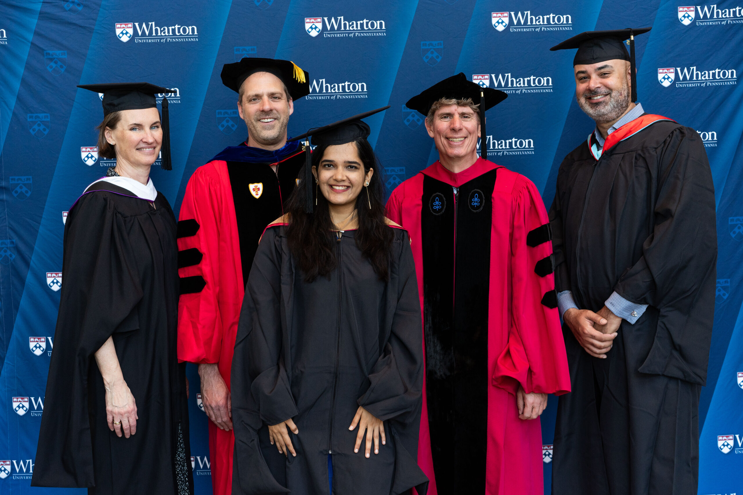 A group of people in academic regalia standing in front of a Wharton School backdrop.