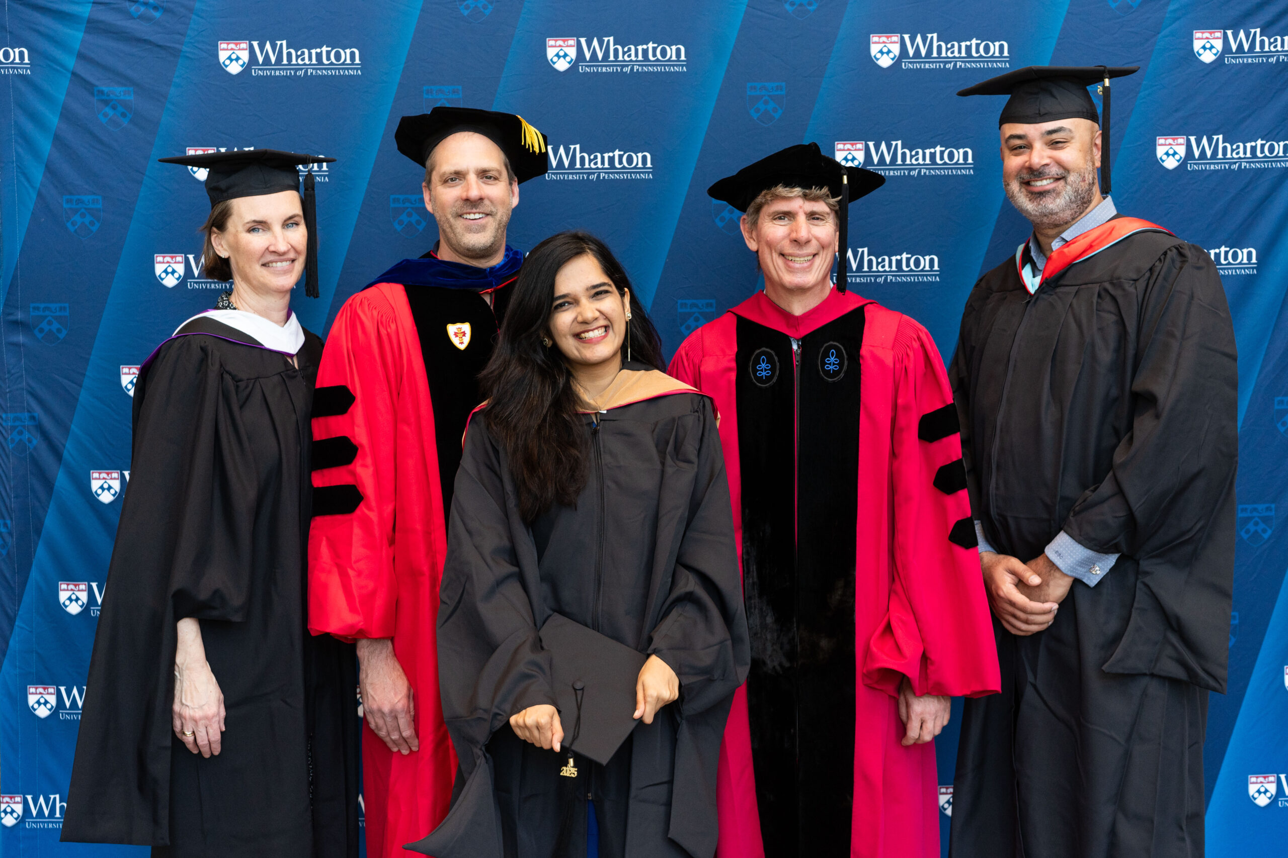A group of people dressed in academic regalia, including caps and gowns, posing in front of a Wharton School, University of Pennsylvania backdrop, suggesting a graduation or academic event.