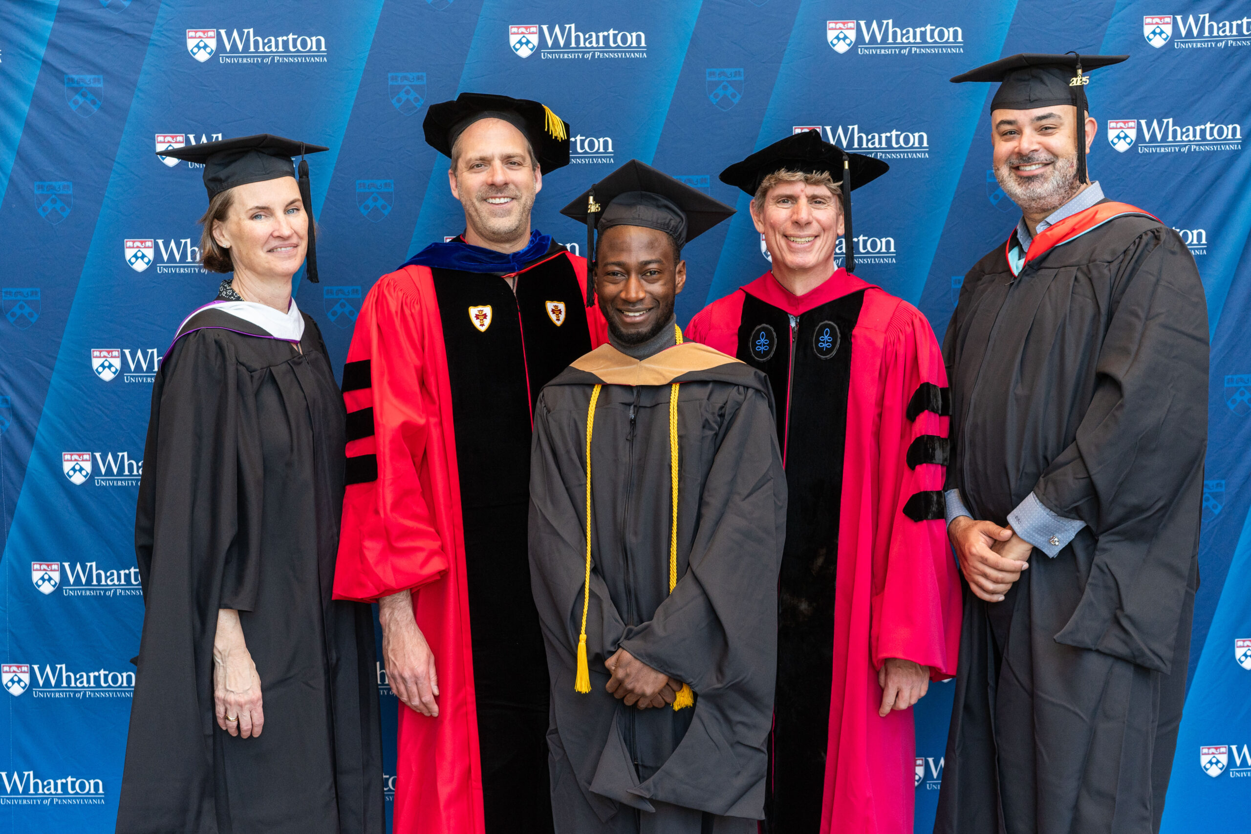 A group of people in academic regalia, including caps and gowns, posing against a Wharton School backdrop, indicating a graduation or academic event.