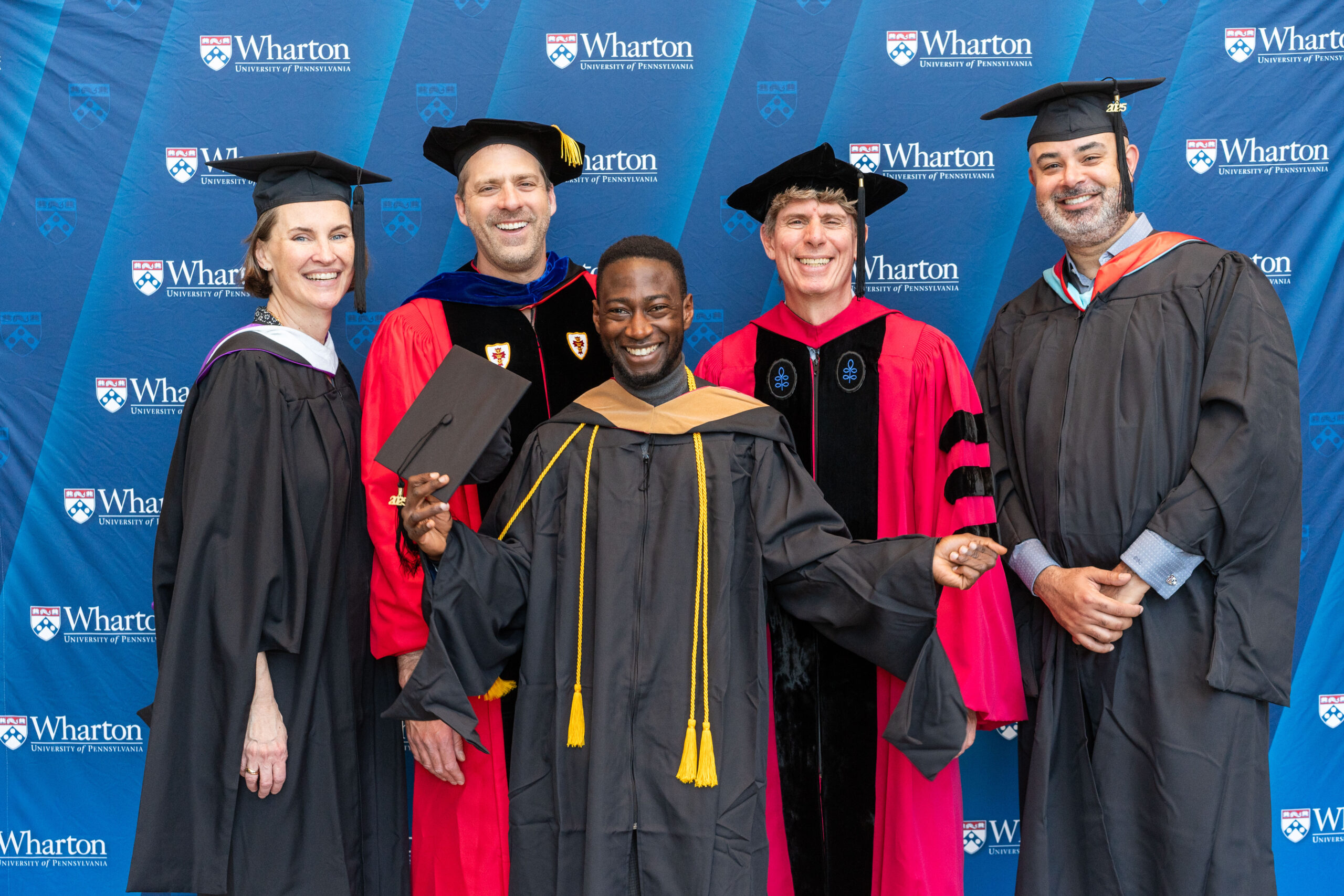 A small group of people wearing graduation attire, including gowns and caps, standing together in front of a "Wharton University of Pennsylvania" backdrop. A person in the center is holding a diploma cover and smiling.