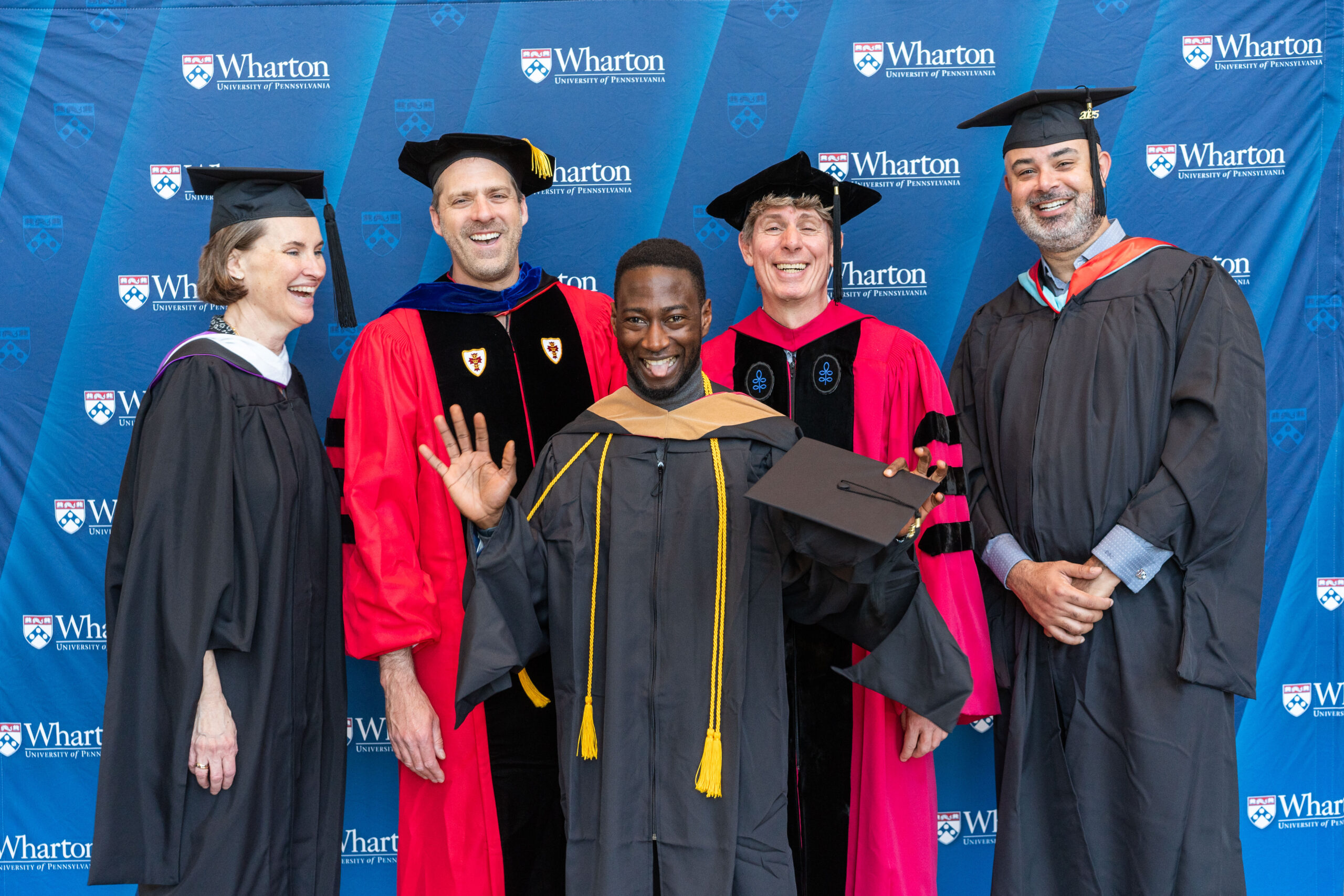 A graduate in a cap and gown poses with faculty members in academic regalia in front of a Wharton University of Pennsylvania banner, smiling and celebrating.