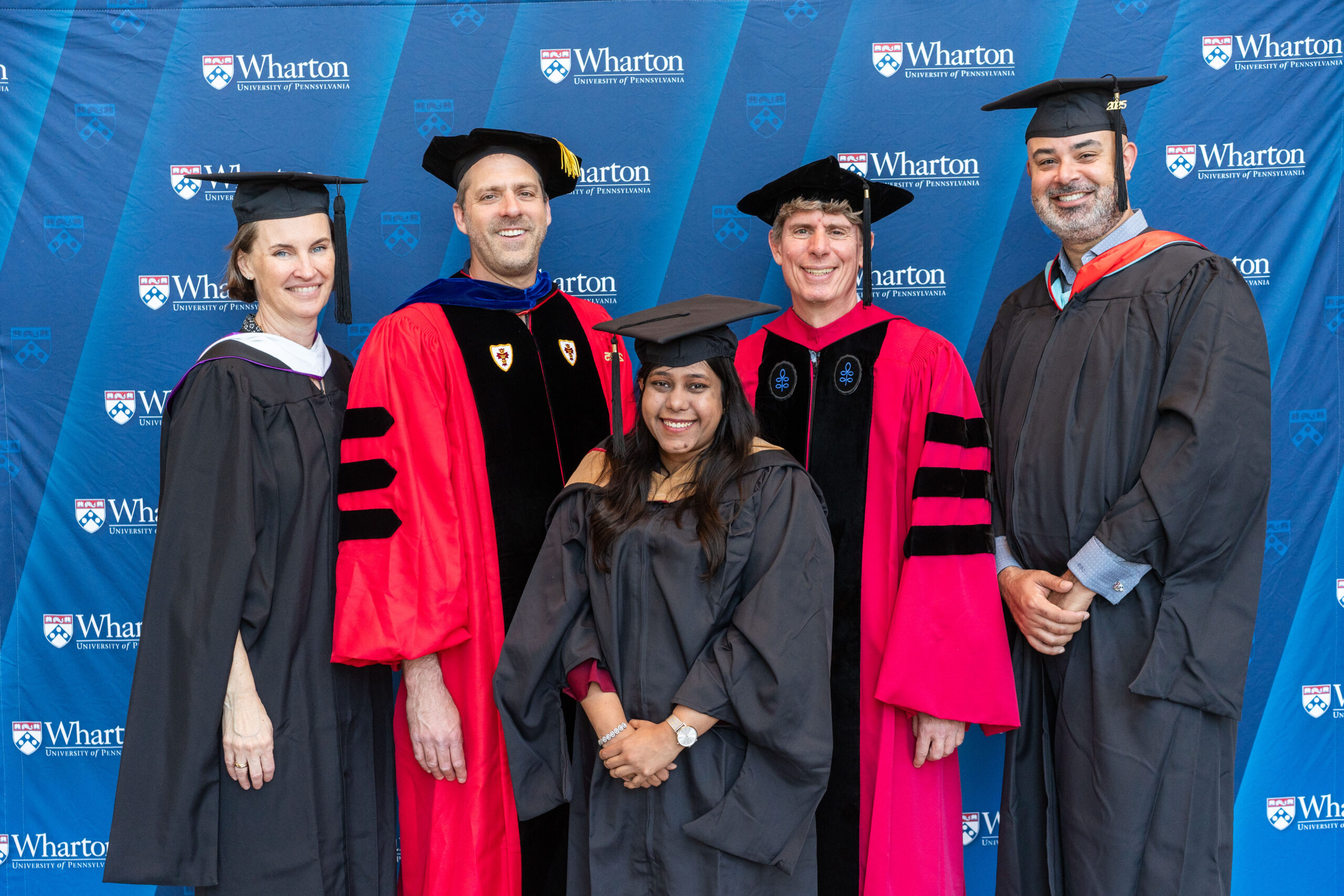 A small group of people in academic regalia, including a graduate in the center, posing in front of a Wharton School of the University of Pennsylvania backdrop.