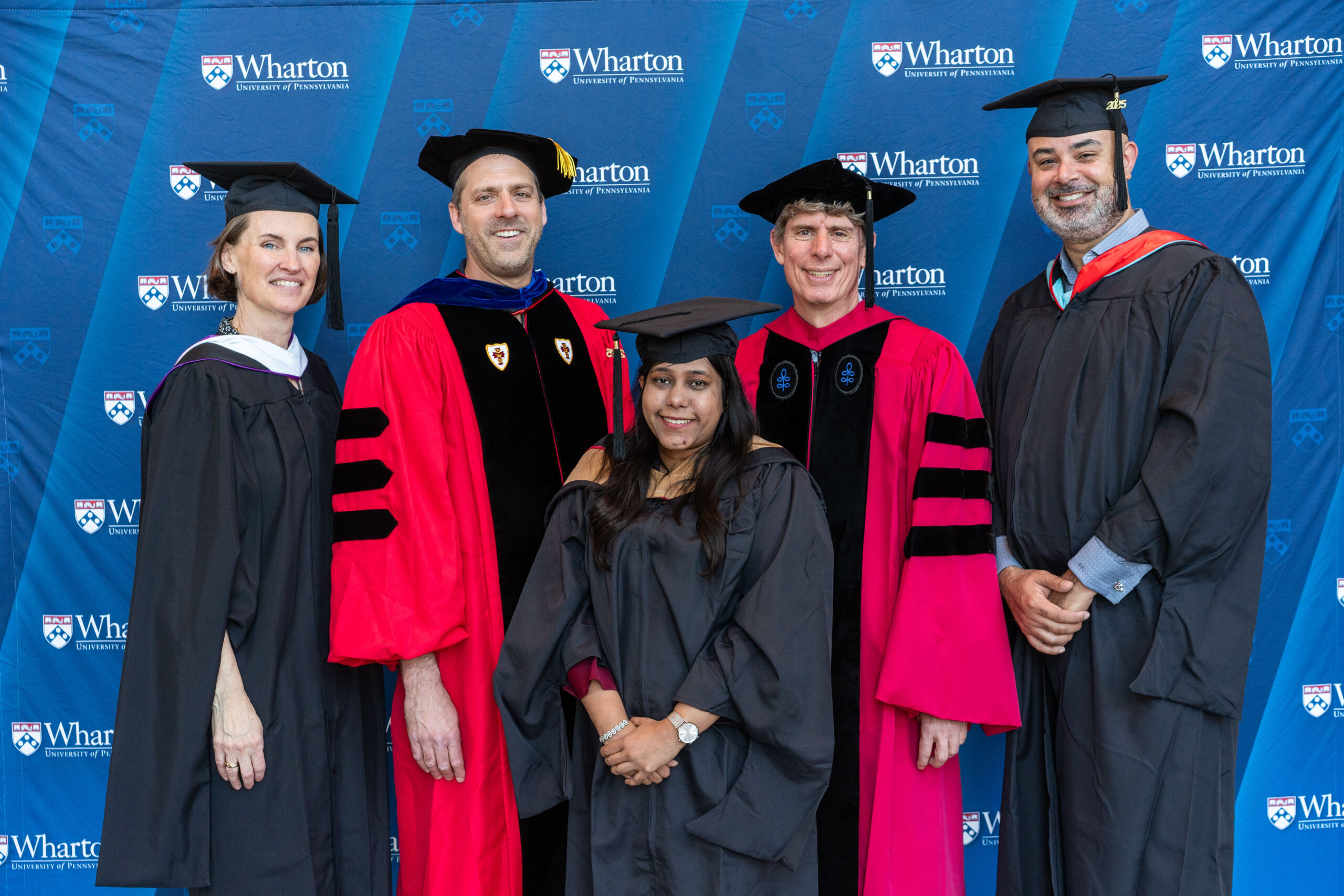 A group of people in academic regalia pose in front of a Wharton School backdrop at a graduation event.