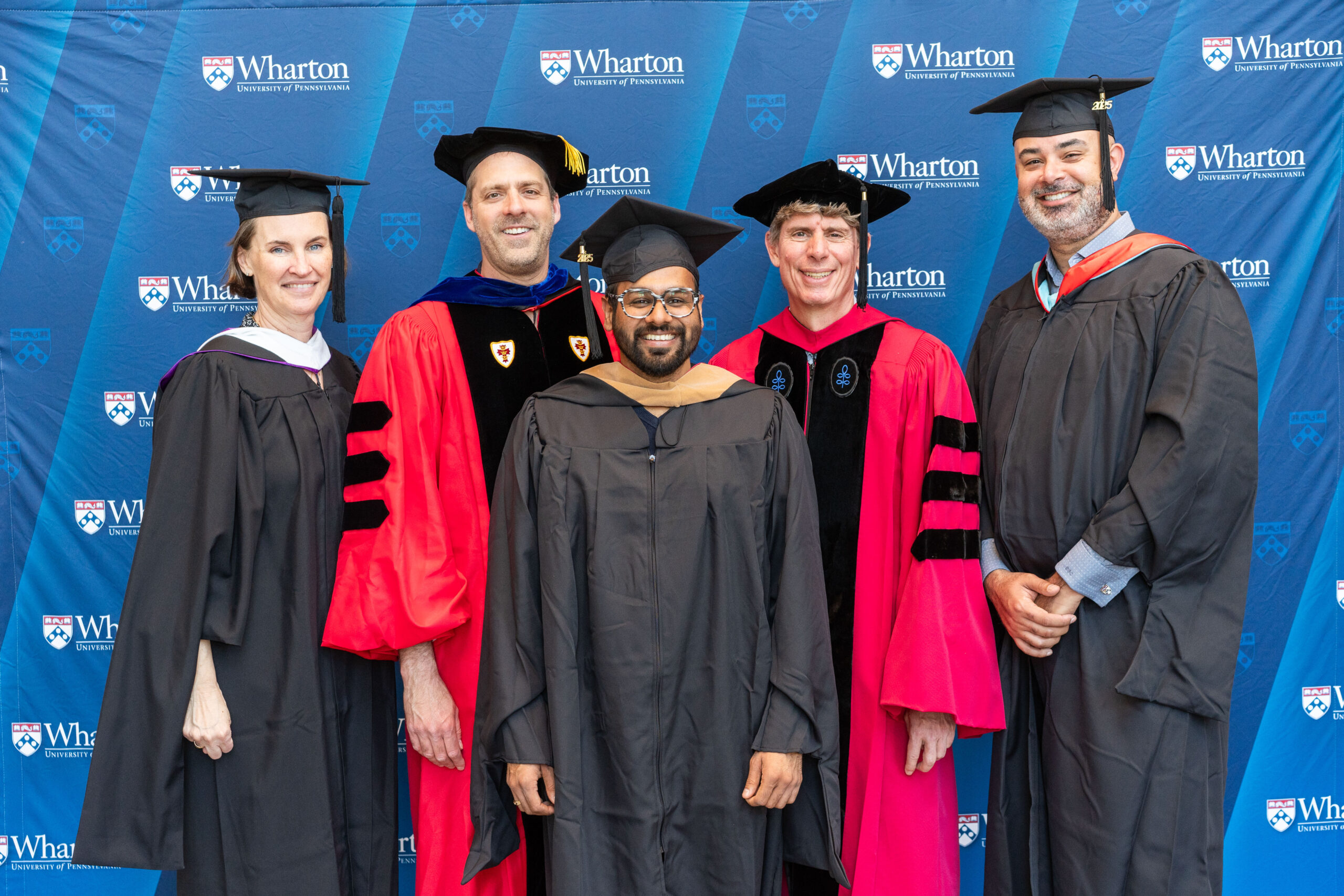 A group of people in academic regalia standing in front of a backdrop with the Wharton School logo, suggesting a graduation or academic event.