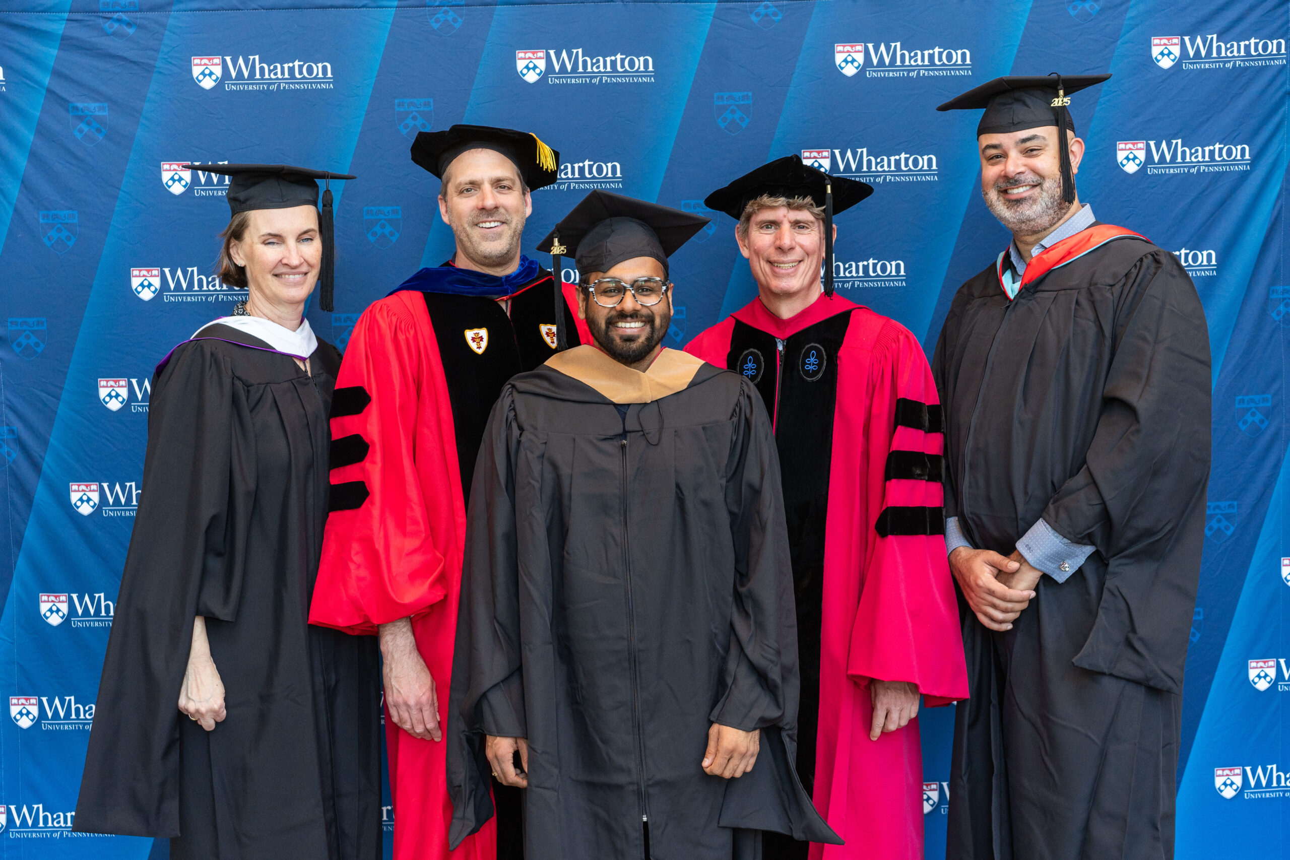 A group of people in academic regalia, including caps and gowns, standing in front of a Wharton University of Pennsylvania backdrop.