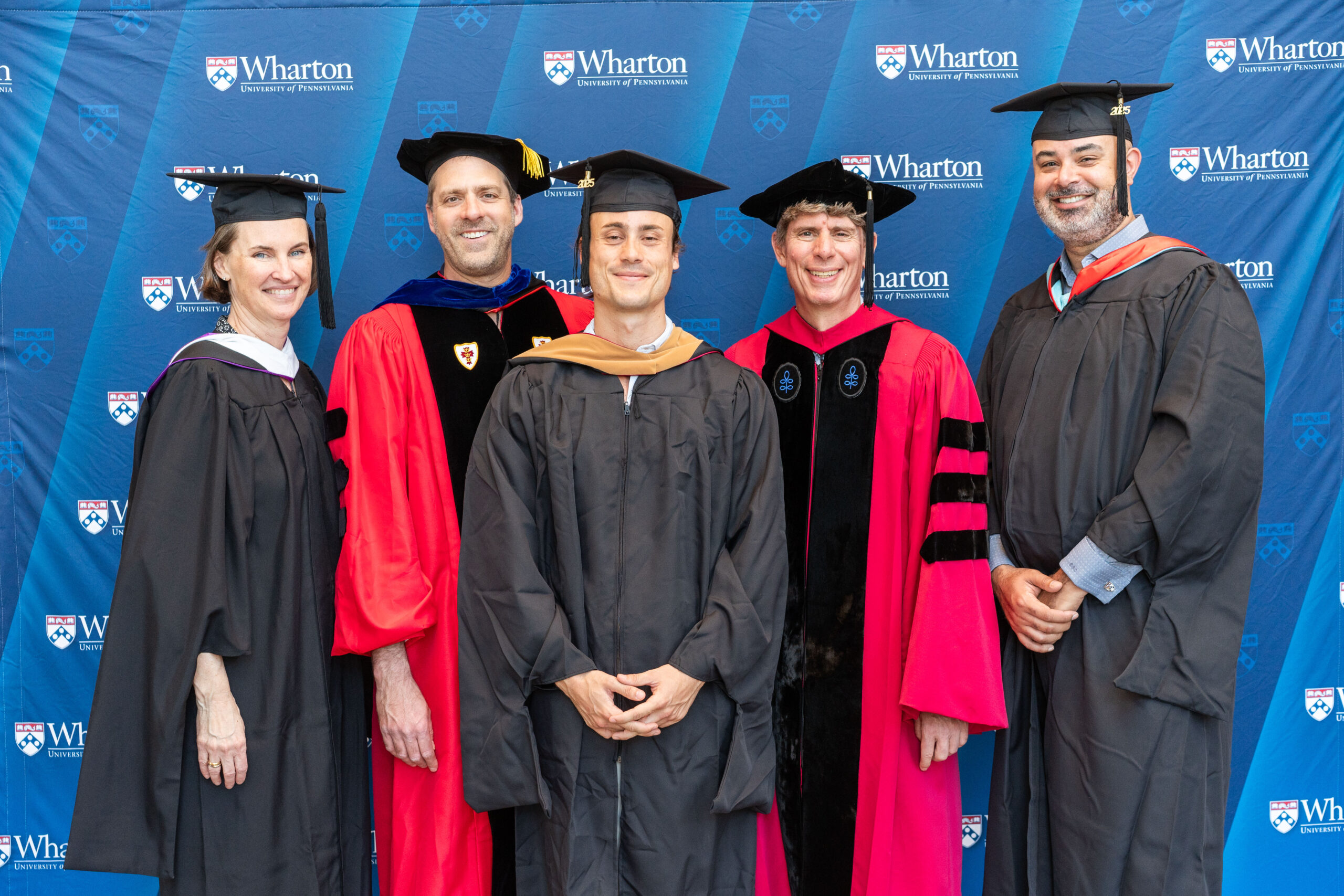 A group of people in academic regalia, including caps and gowns, posing in front of a backdrop with "Wharton University of Pennsylvania" logos, suggesting a graduation or academic event.