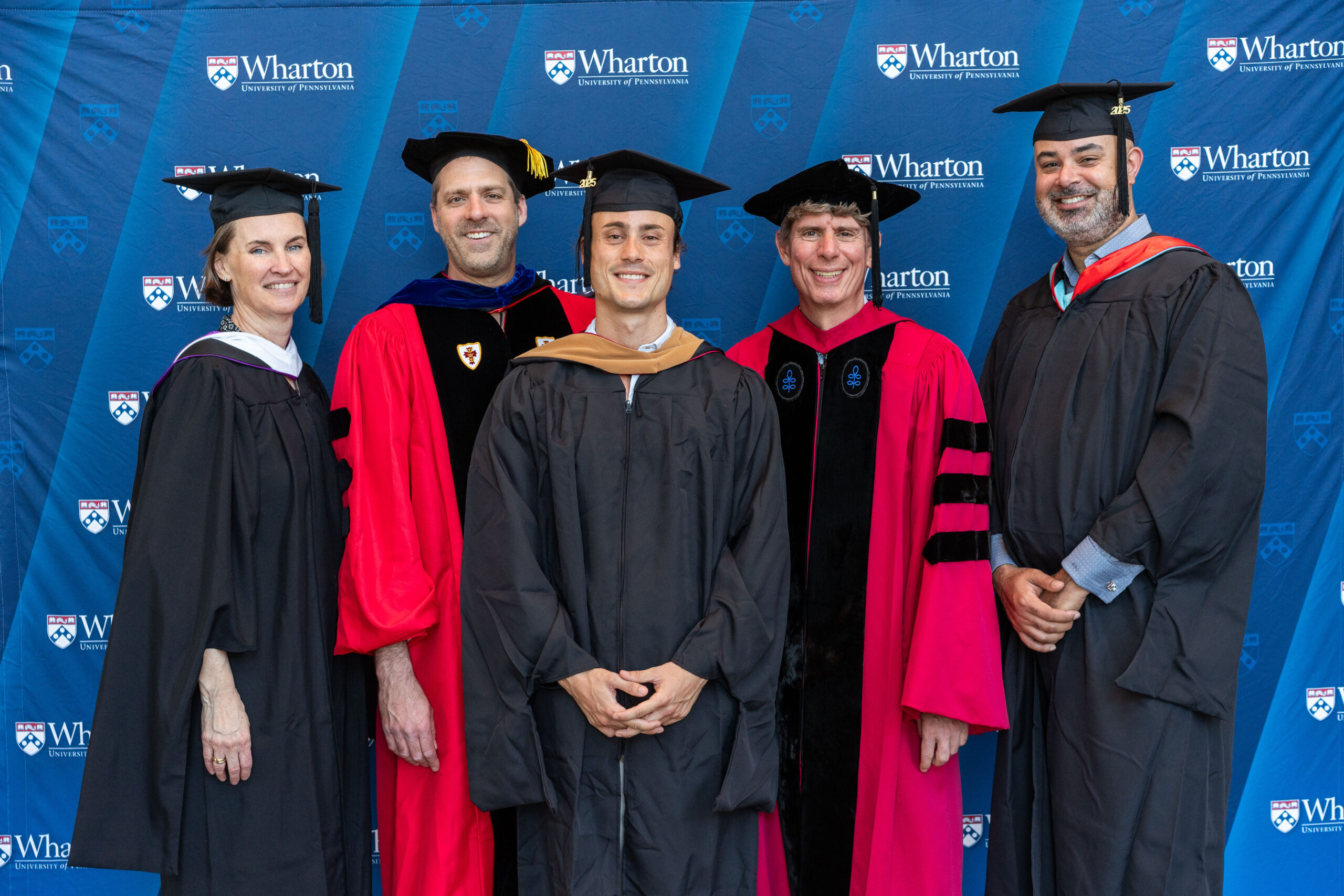 A small group wearing academic regalia, smiling in front of a Wharton School backdrop, indicating a graduation or academic event.