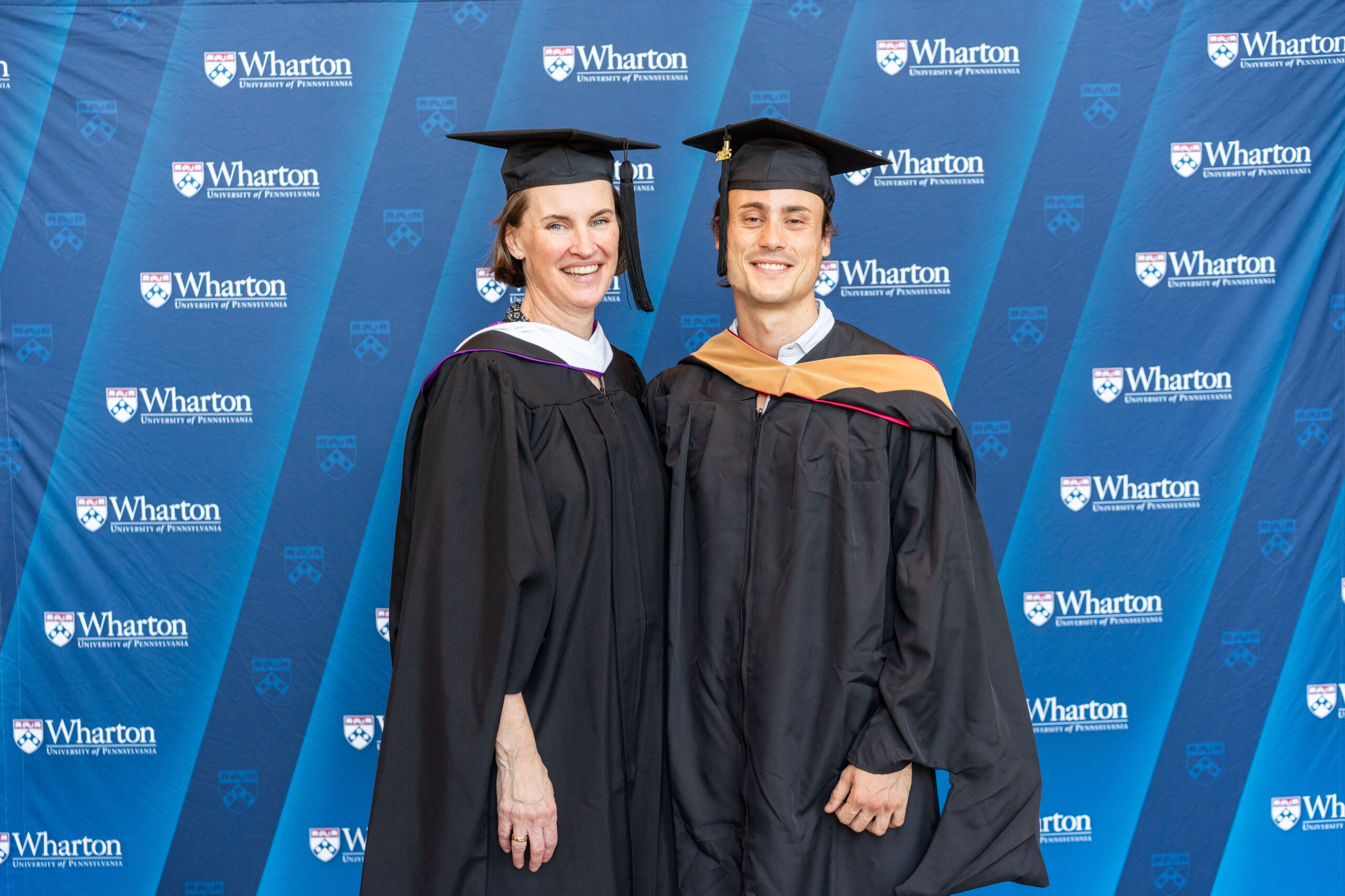 A couple wearing graduation caps and gowns stands in front of a backdrop with the Wharton School logo, smiling at the camera.