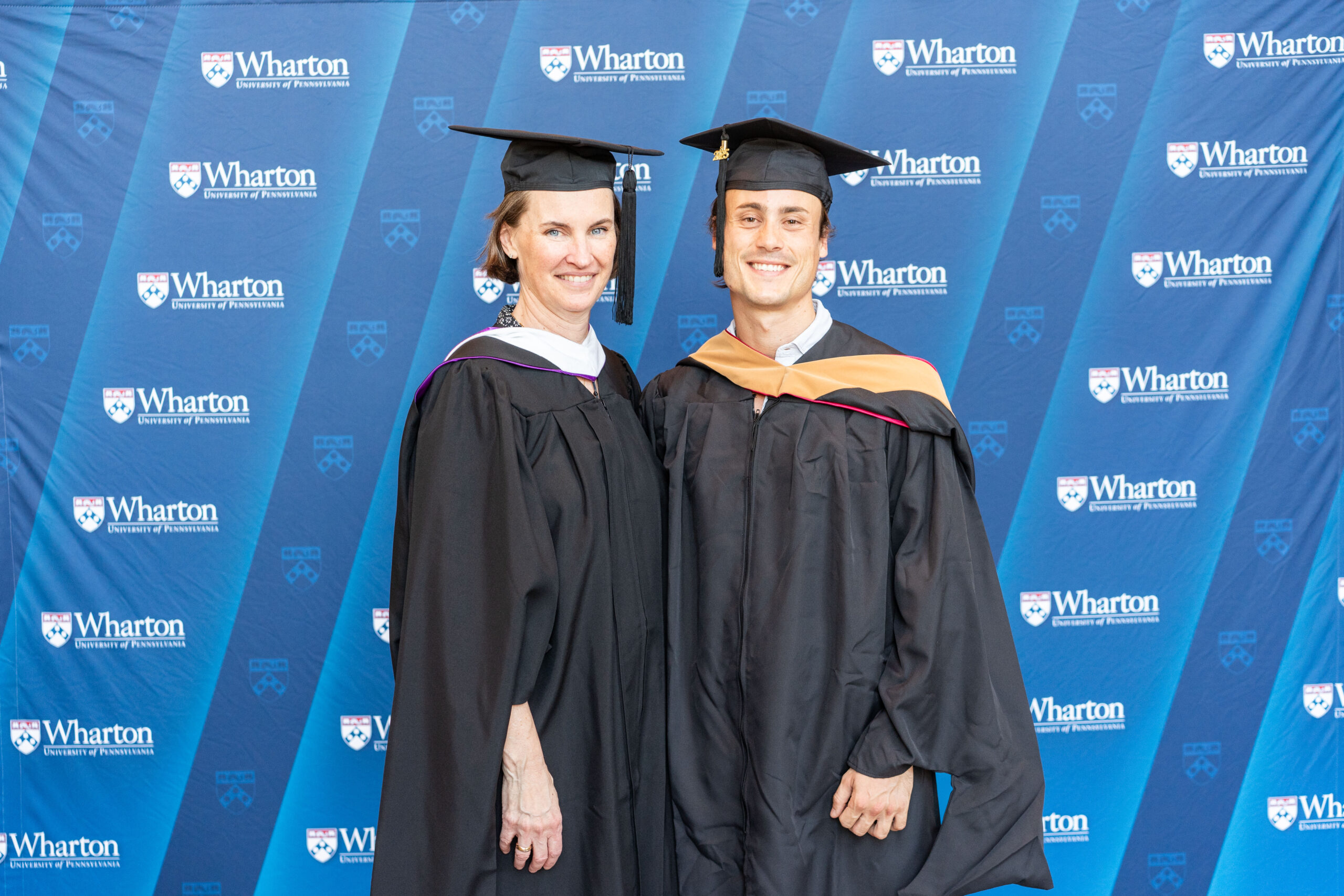 Two individuals wearing graduation caps and gowns stand in front of a backdrop featuring the Wharton School logo.