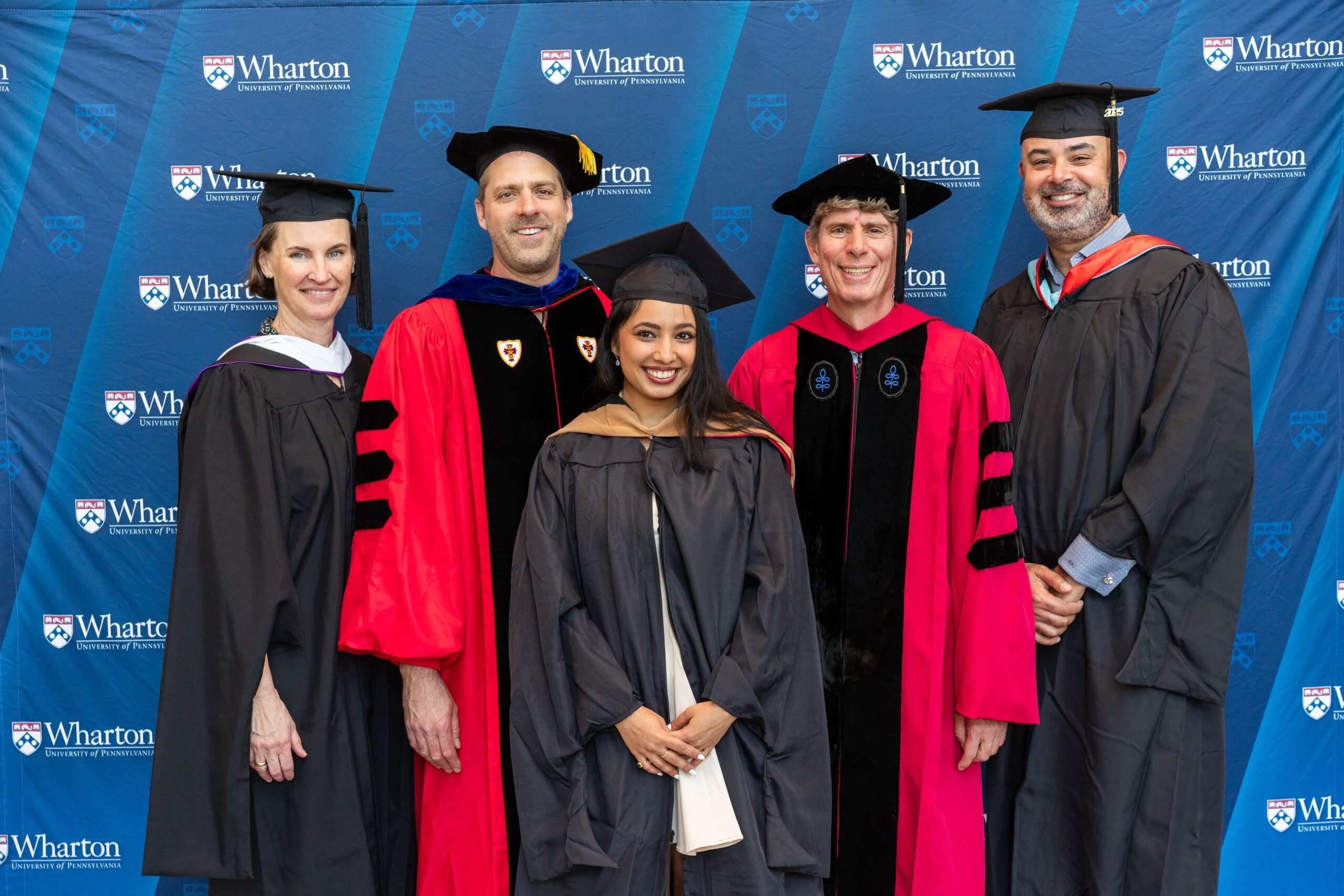 A small group of people in academic regalia pose in front of a backdrop featuring "Wharton University of Pennsylvania" logos.