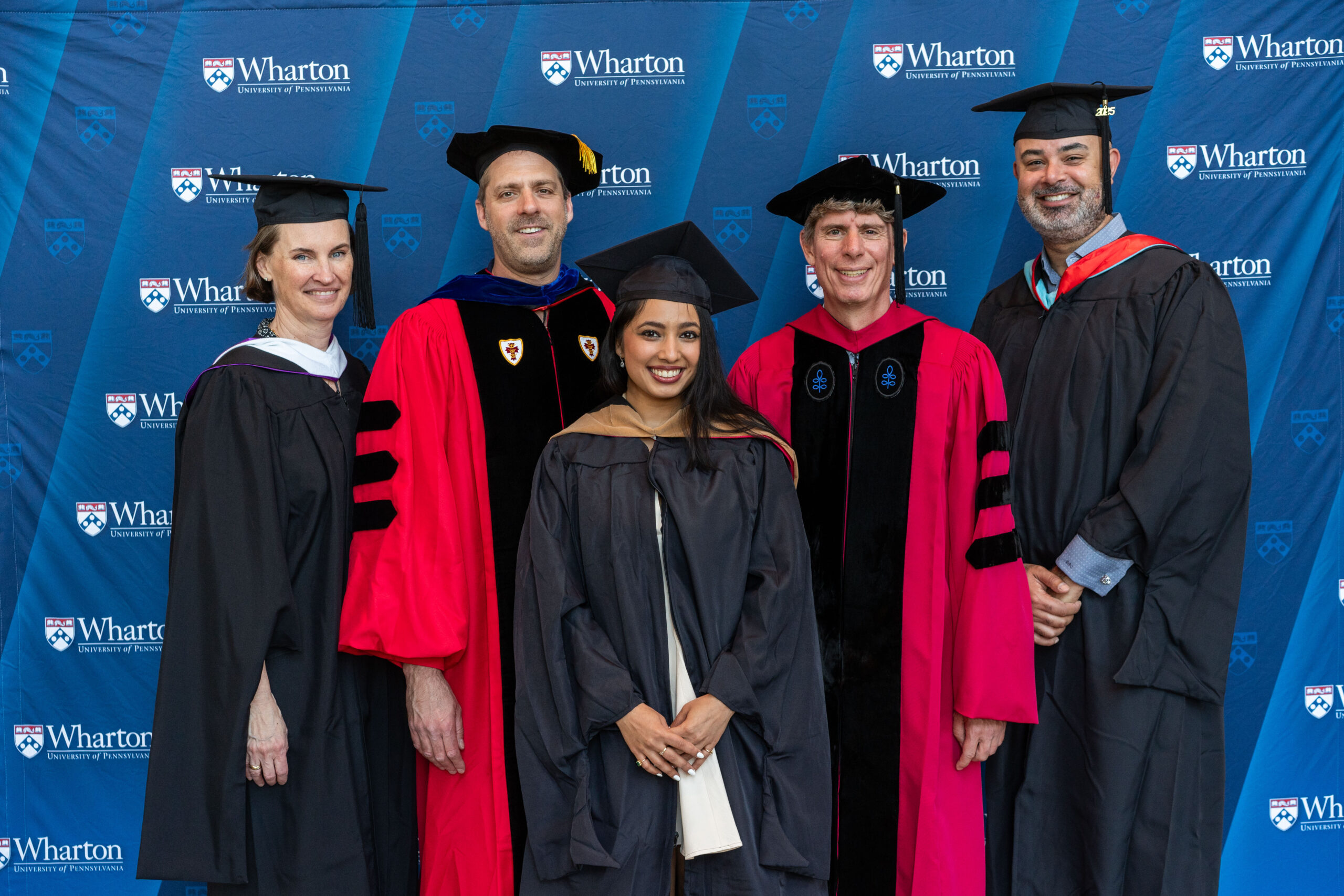 A group of individuals in graduation attire stands in front of a backdrop with "Wharton University of Pennsylvania" logos. Some are wearing academic robes with caps and others are in more formal academic regalia.