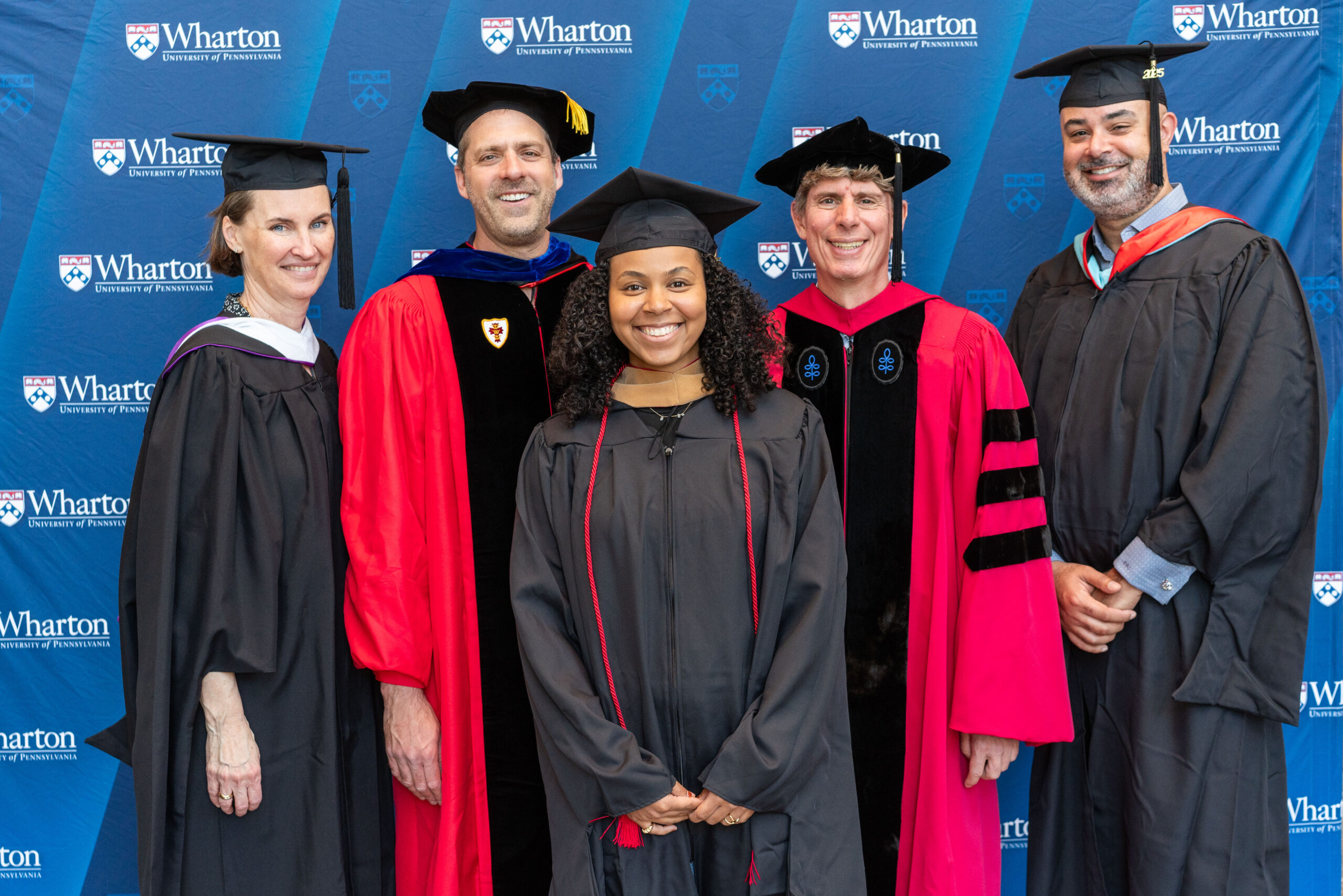 A group of people in academic regalia, including graduation robes and caps, pose in front of a Wharton University of Pennsylvania banner.
