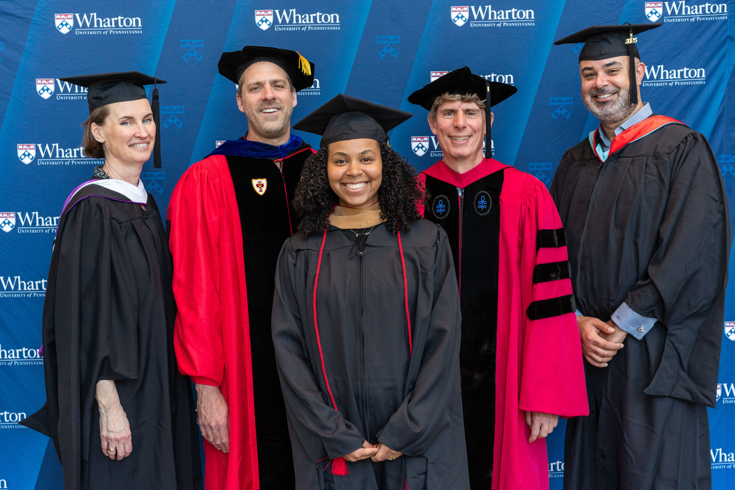 A group of people in graduation attire, including caps and gowns, standing in front of a Wharton University of Pennsylvania backdrop.