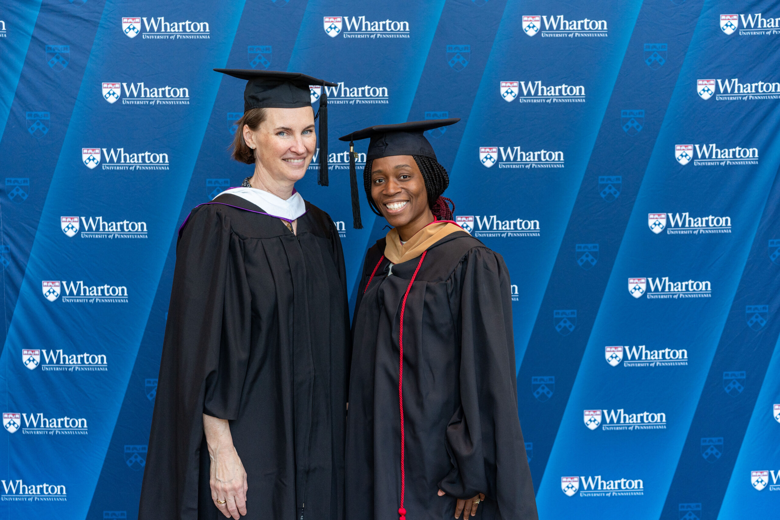 A couple of people in graduation gowns and caps stand in front of a blue backdrop with "Wharton University of Pennsylvania" logos.