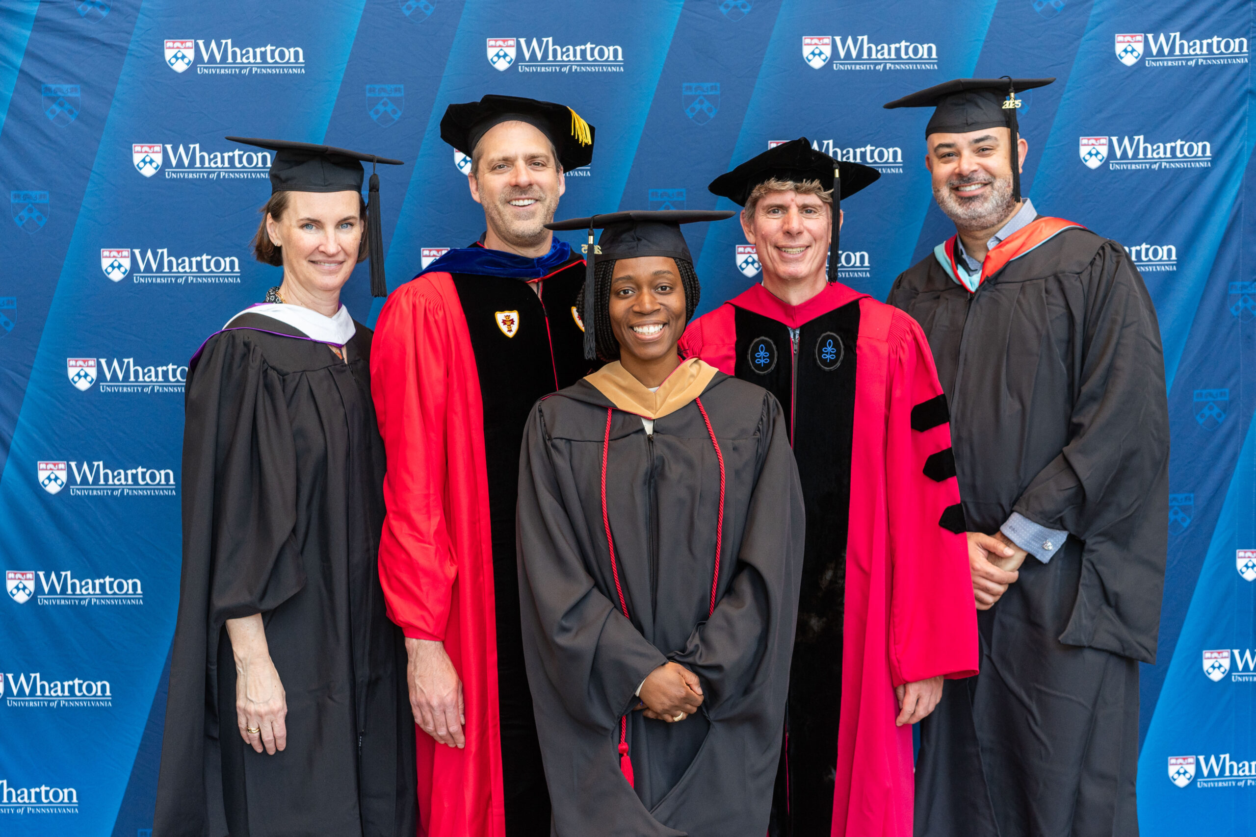 A small group of people in academic regalia, including caps and gowns, posing for a photo in front of a Wharton School backdrop.