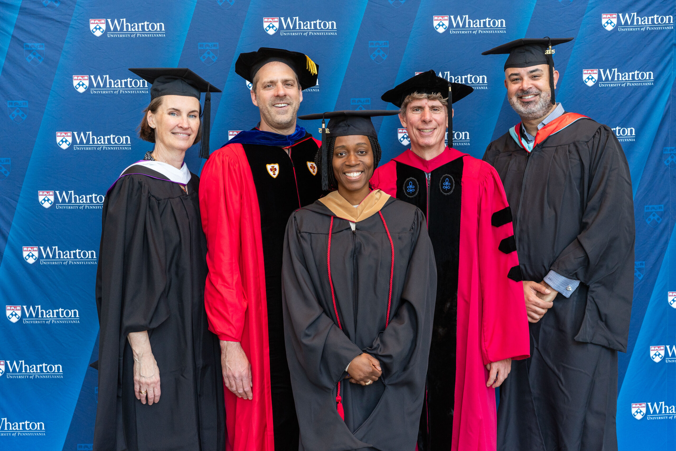 A group of individuals in academic regalia, including graduation caps and gowns, standing in front of a Wharton School of the University of Pennsylvania backdrop, suggesting a graduation or academic event.
