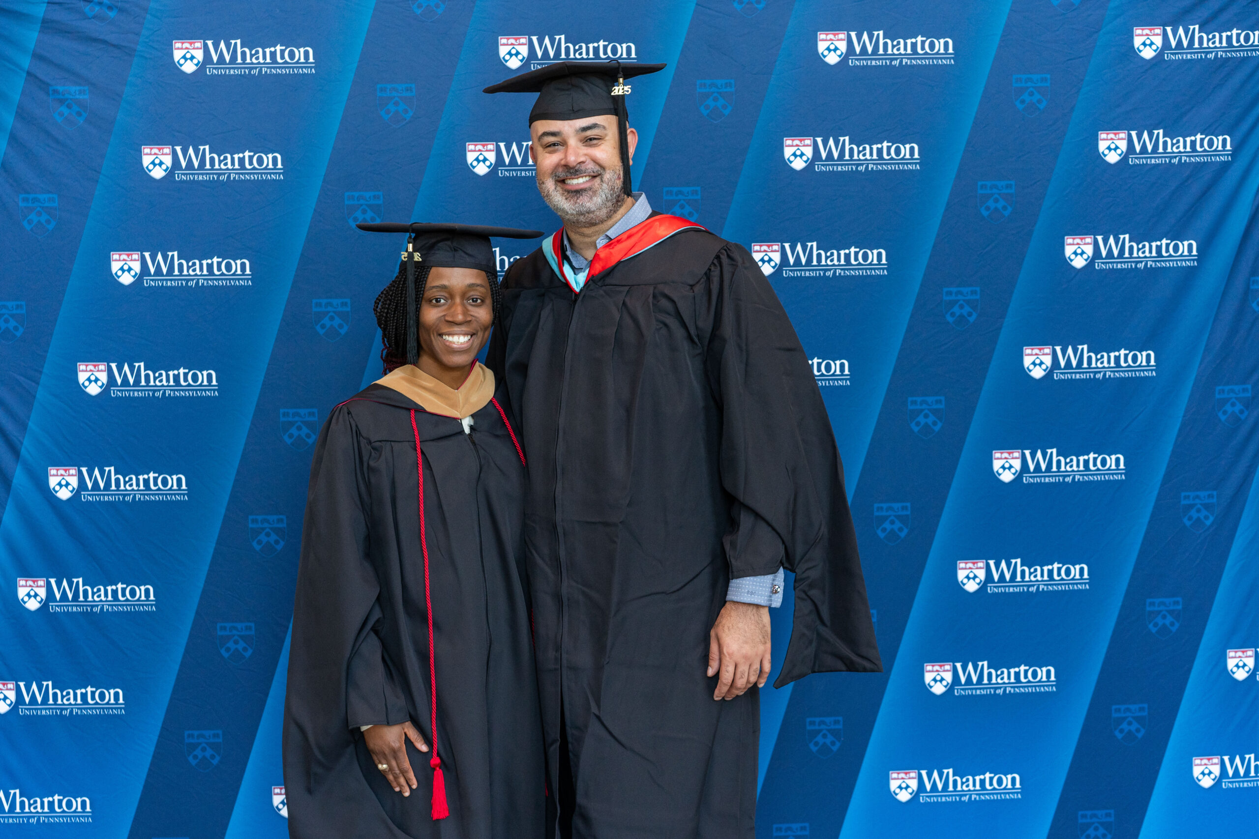 A couple wearing graduation caps and gowns stands in front of a backdrop with the Wharton School logo, indicating a graduation ceremony.