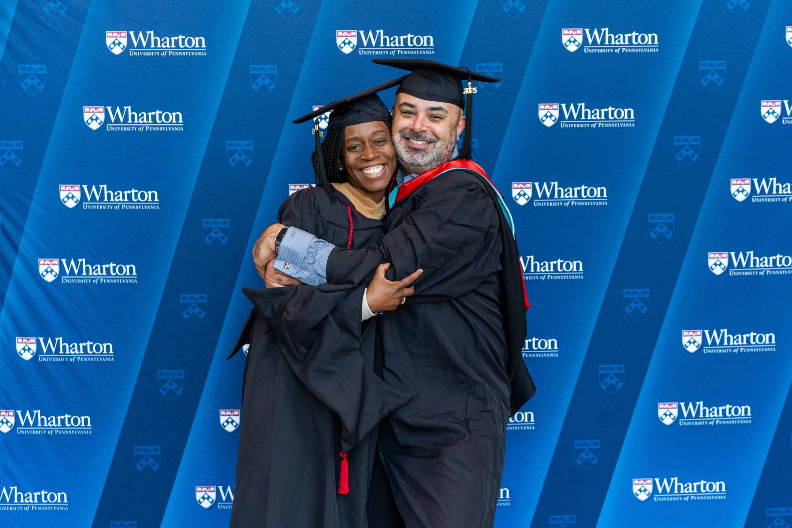 A couple of graduates in caps and gowns embrace and smile in front of a Wharton School of the University of Pennsylvania backdrop.
